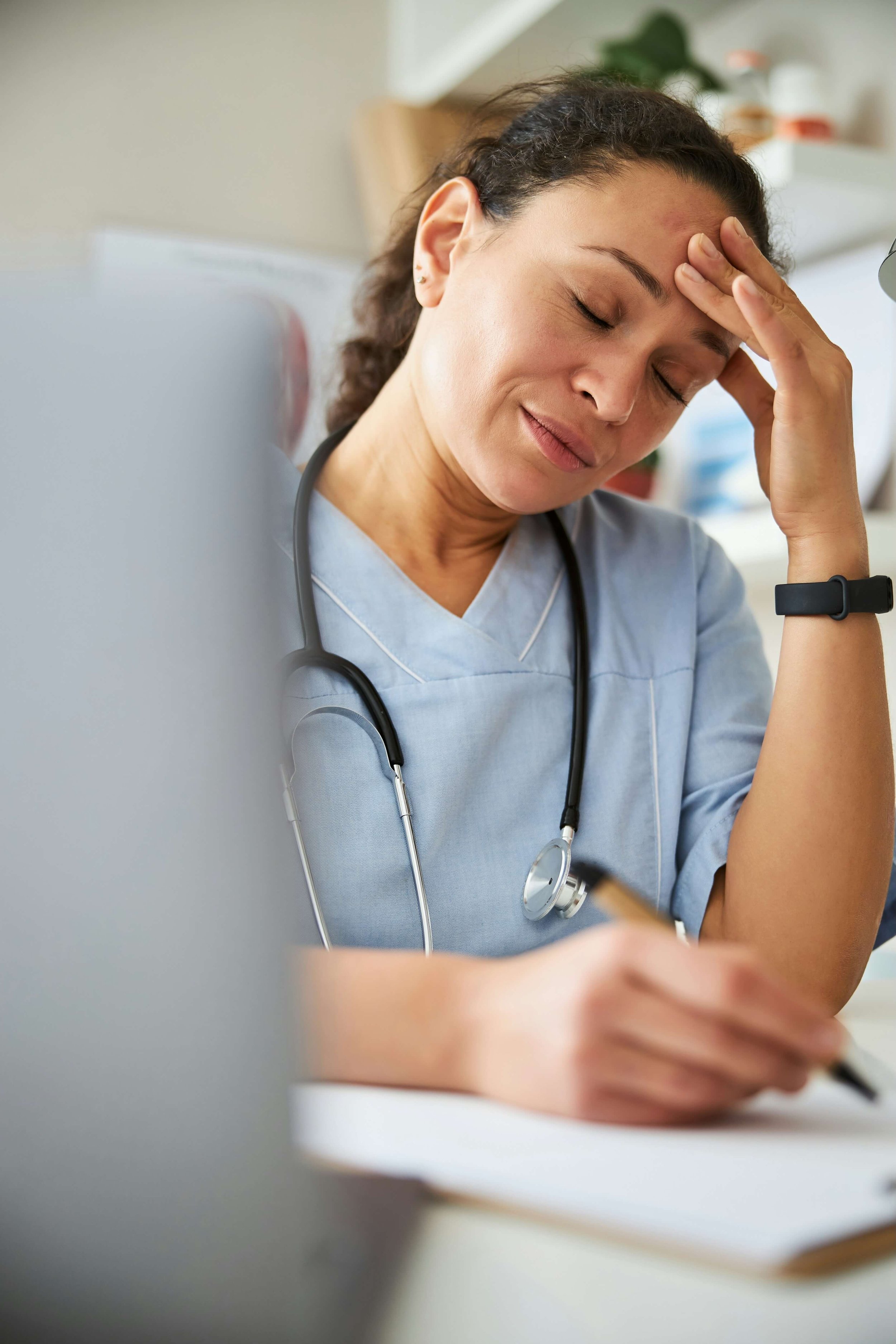 A female healthcare professional with a stethoscope around her neck, wearing scrubs, resting her forehead on her hand with eyes closed, appearing stressed or tired, sitting at a desk with a blurred background.