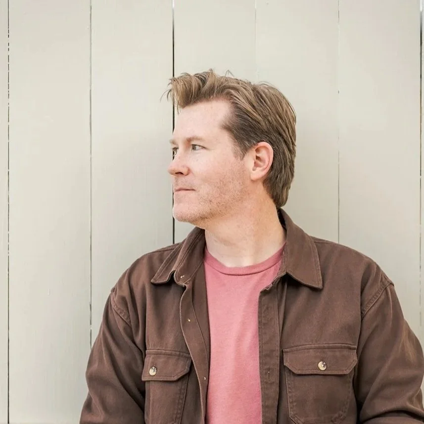 A man with brown hair stands sideways in front of a light-colored wooden wall, looking to his left.
