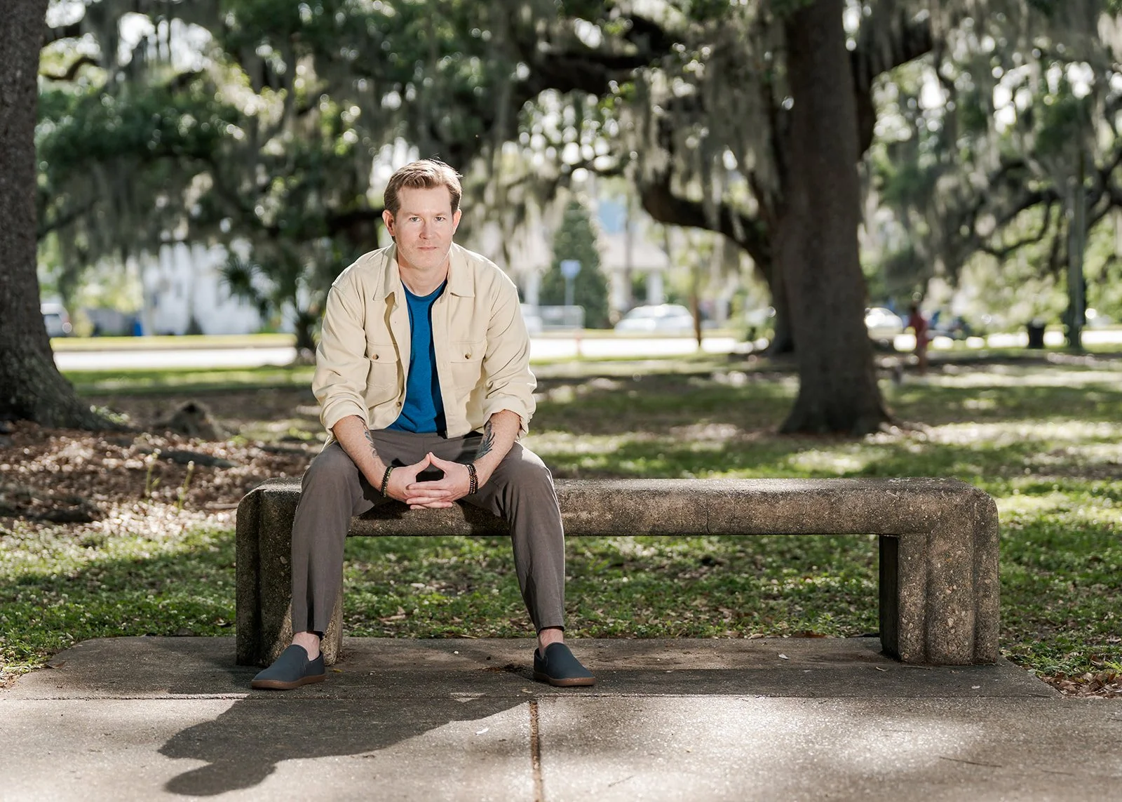 A man sitting on a concrete park bench with large trees and a sunny background.