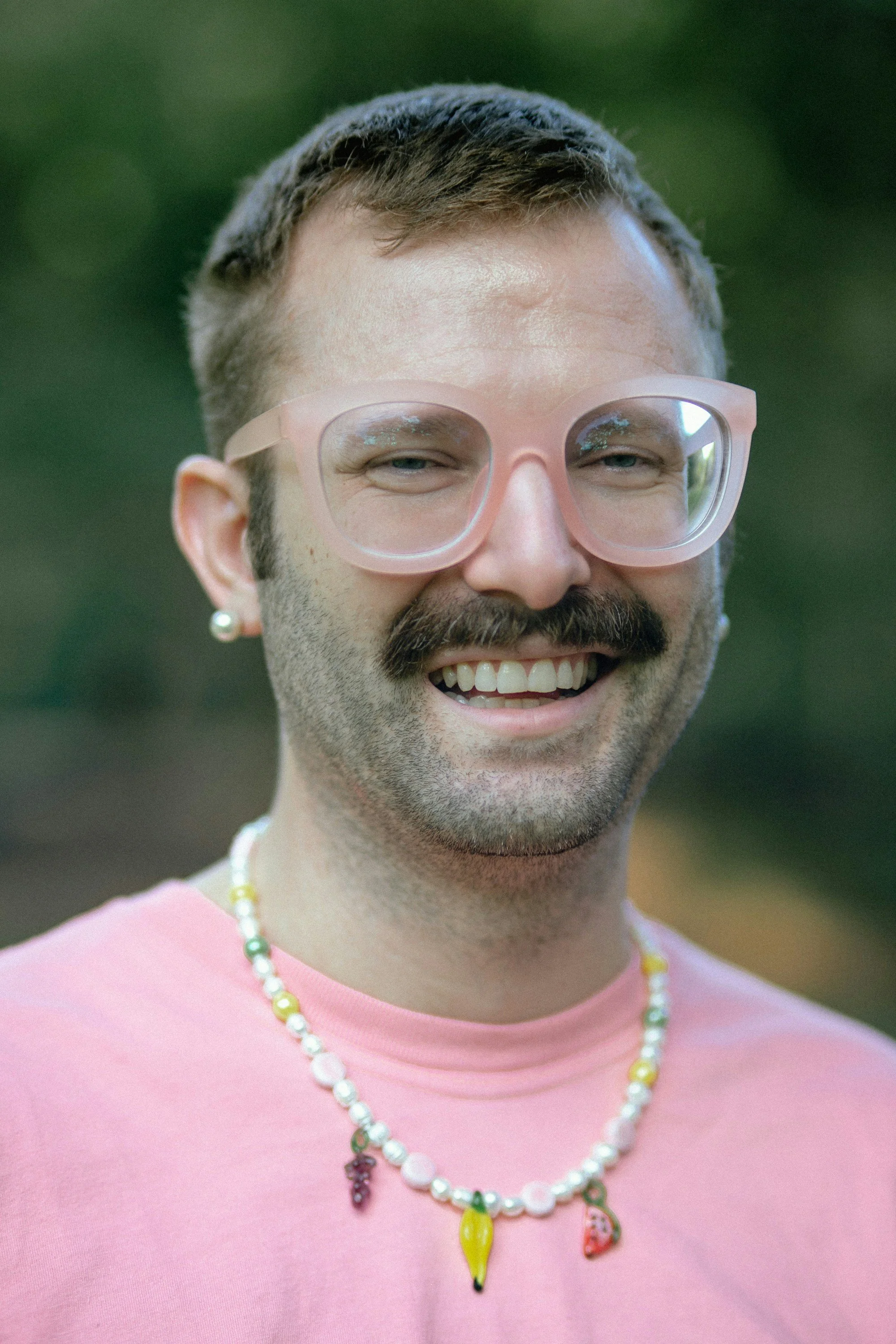 Close-up of a smiling man with a mustache and beard, wearing pink glasses, a pink shirt, and a colorful beaded necklace with charms, outdoors with a blurred green background.