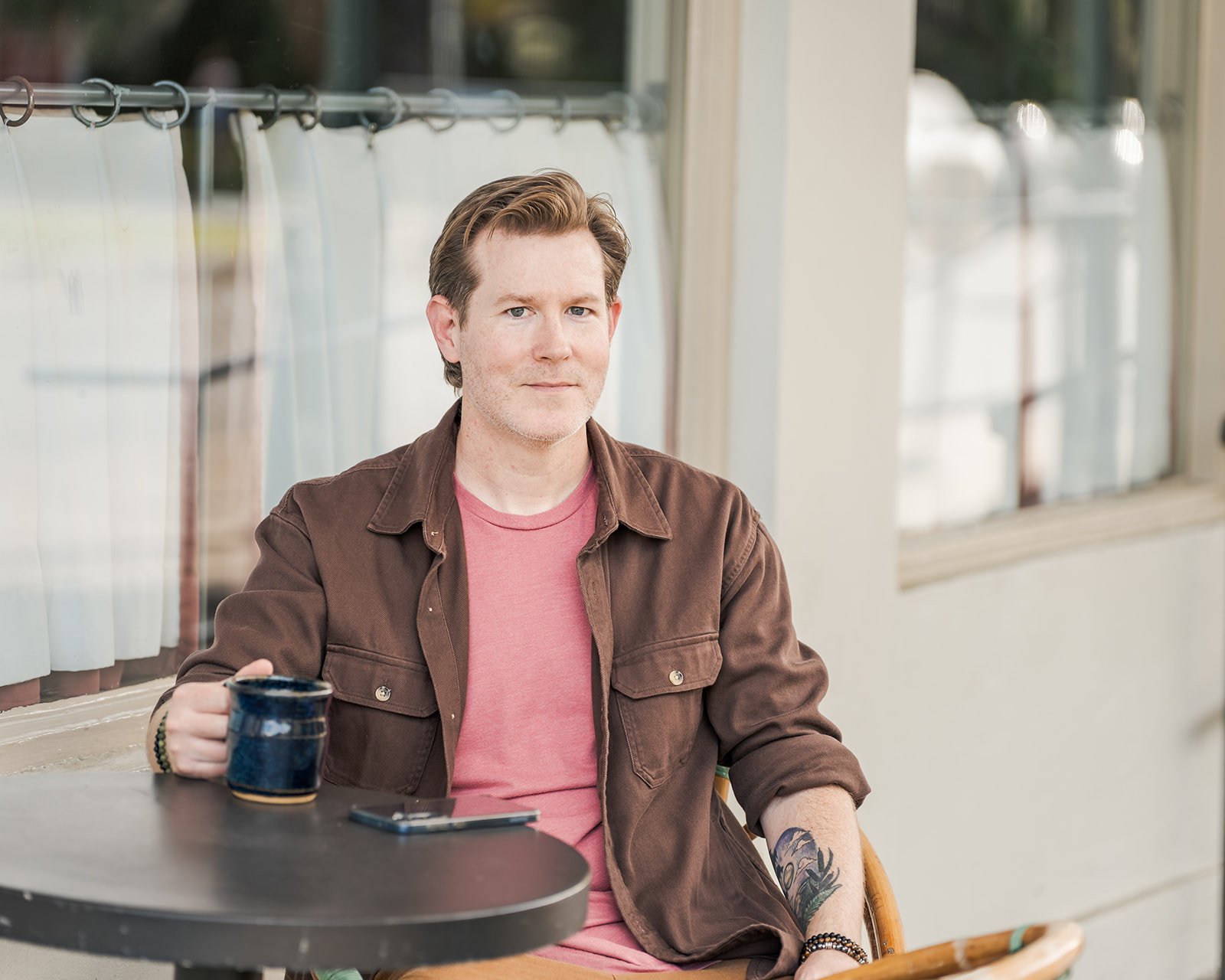 A man with brown hair and tattoos on his arm, sitting at a table holding a mug, with a window and white curtains in the background.