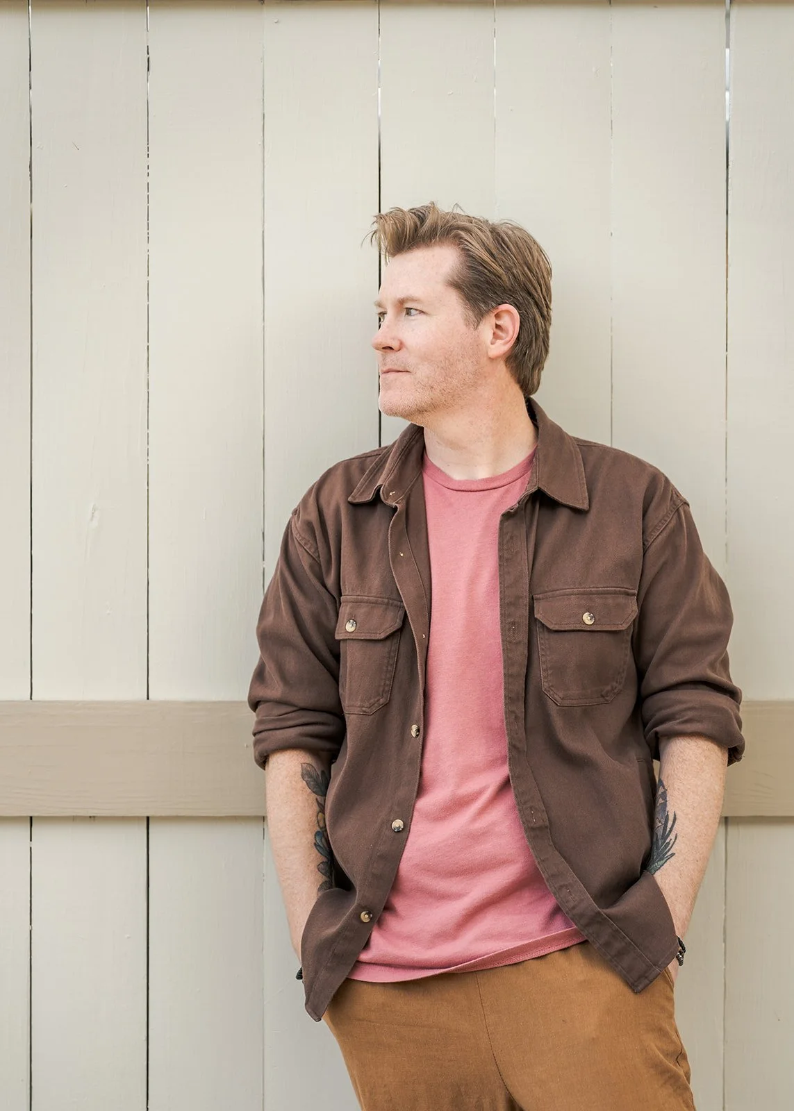 A man with brown hair, wearing a brown jacket over a pink t-shirt and tan pants, leaning against a beige wooden fence with his hands in his pockets.
