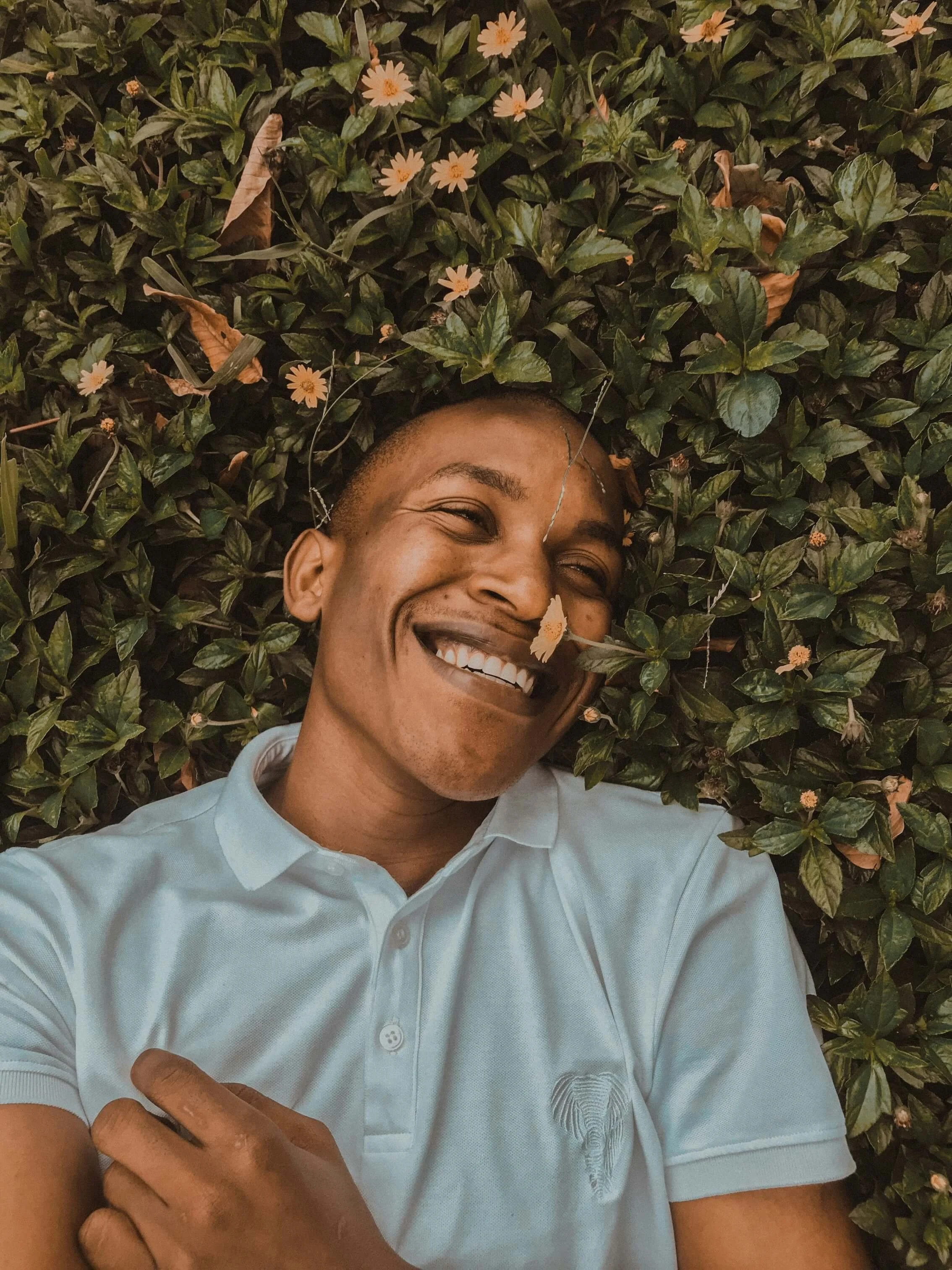 A smiling person lying on a bed of green leaves and small pink flowers, wearing a white shirt.