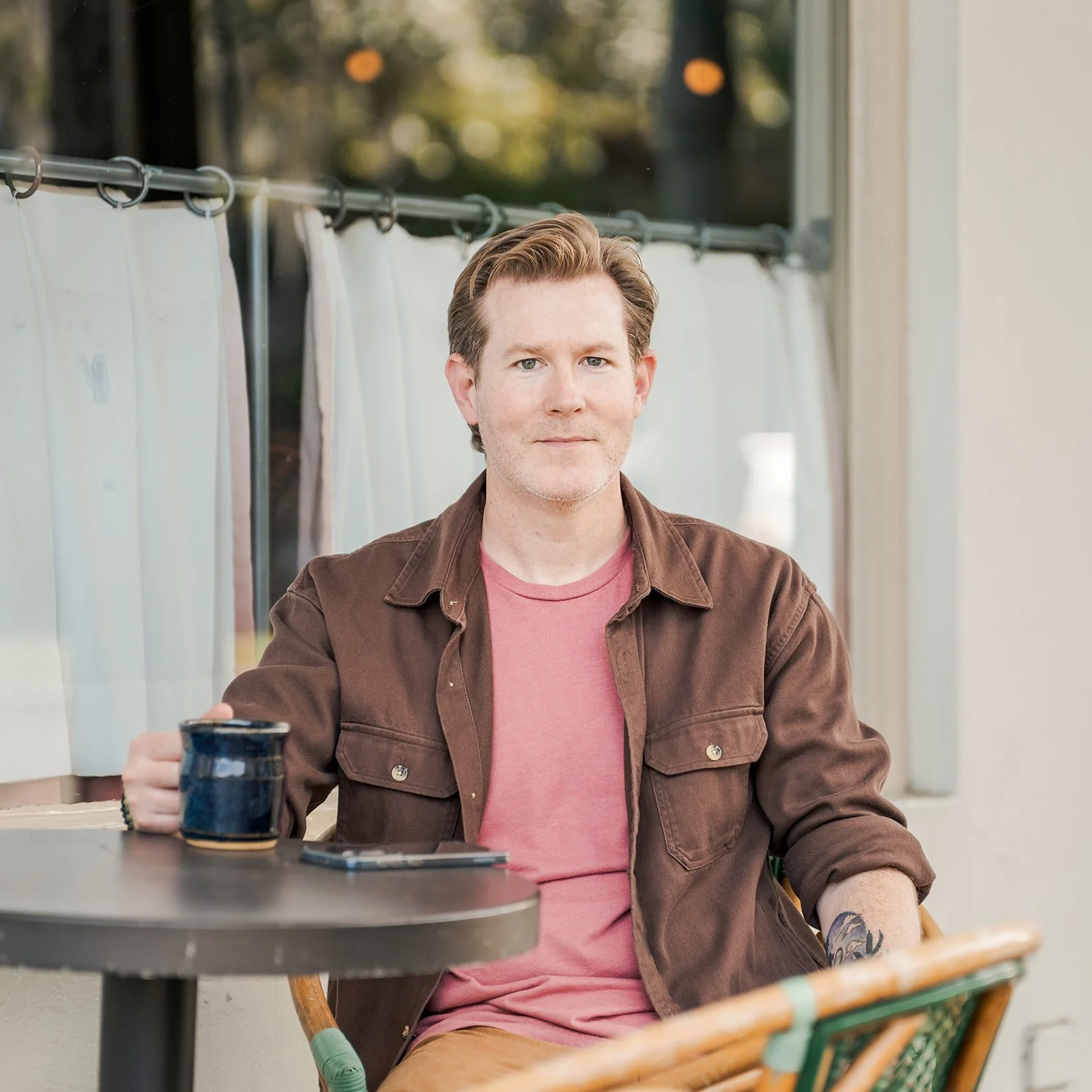 A man with brown hair sitting at a table outdoors, holding a dark blue mug, wearing a pink t-shirt and a brown jacket, with tattoos on his left forearm.