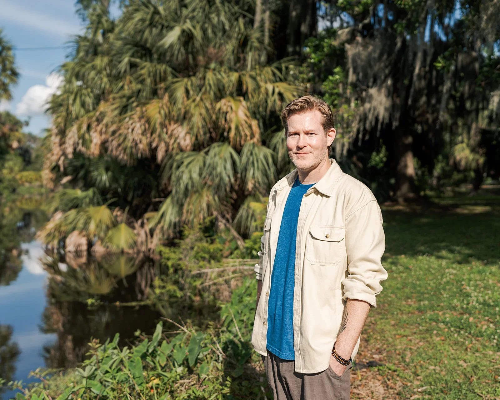 A man with short brown hair wearing a beige jacket, blue t-shirt, and dark pants standing outdoors near a body of water with large leafy trees in the background.