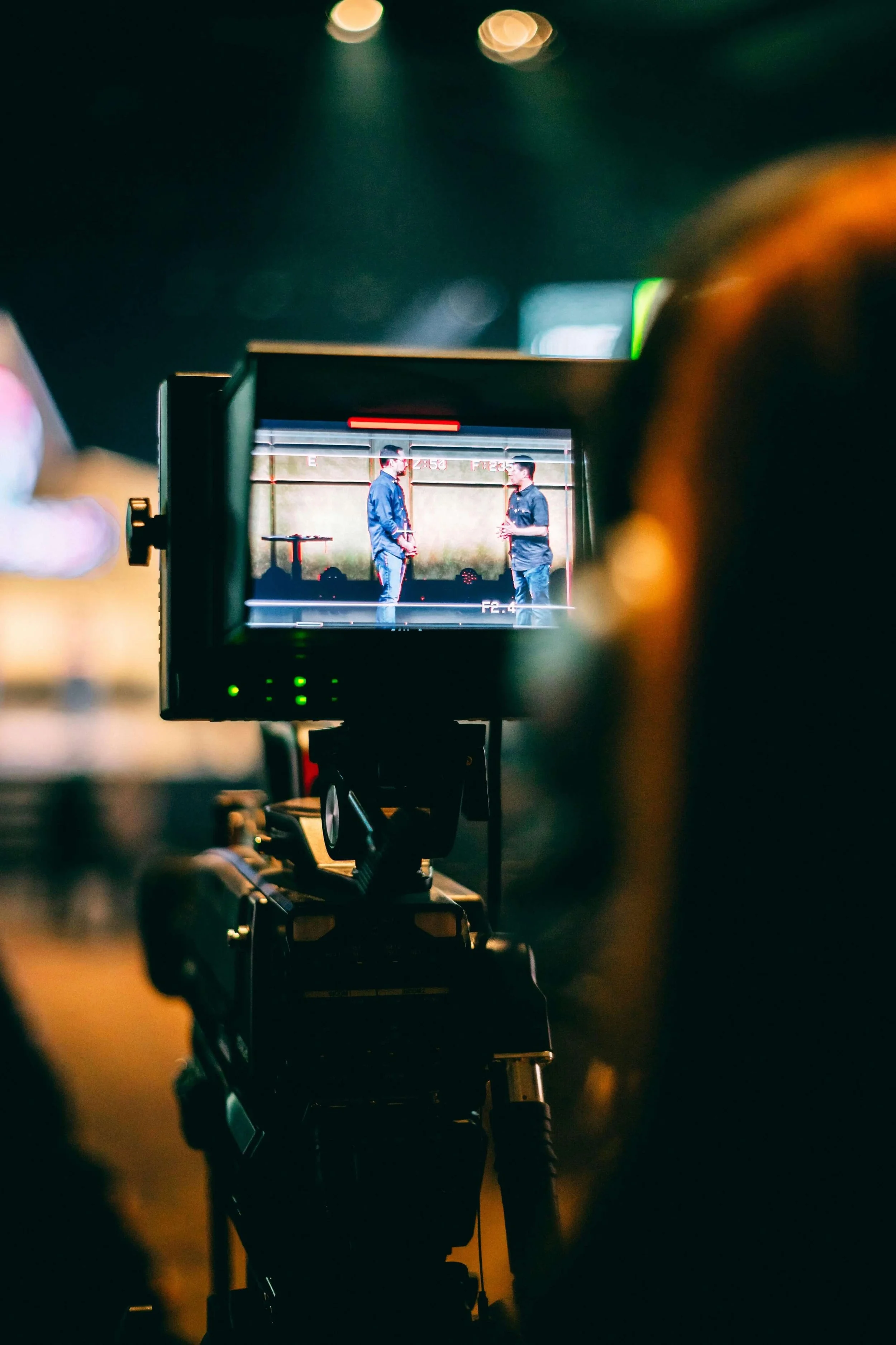 A camera viewfinder displaying two men in conversation, standing on a stage or in a studio environment with a gradient background.
