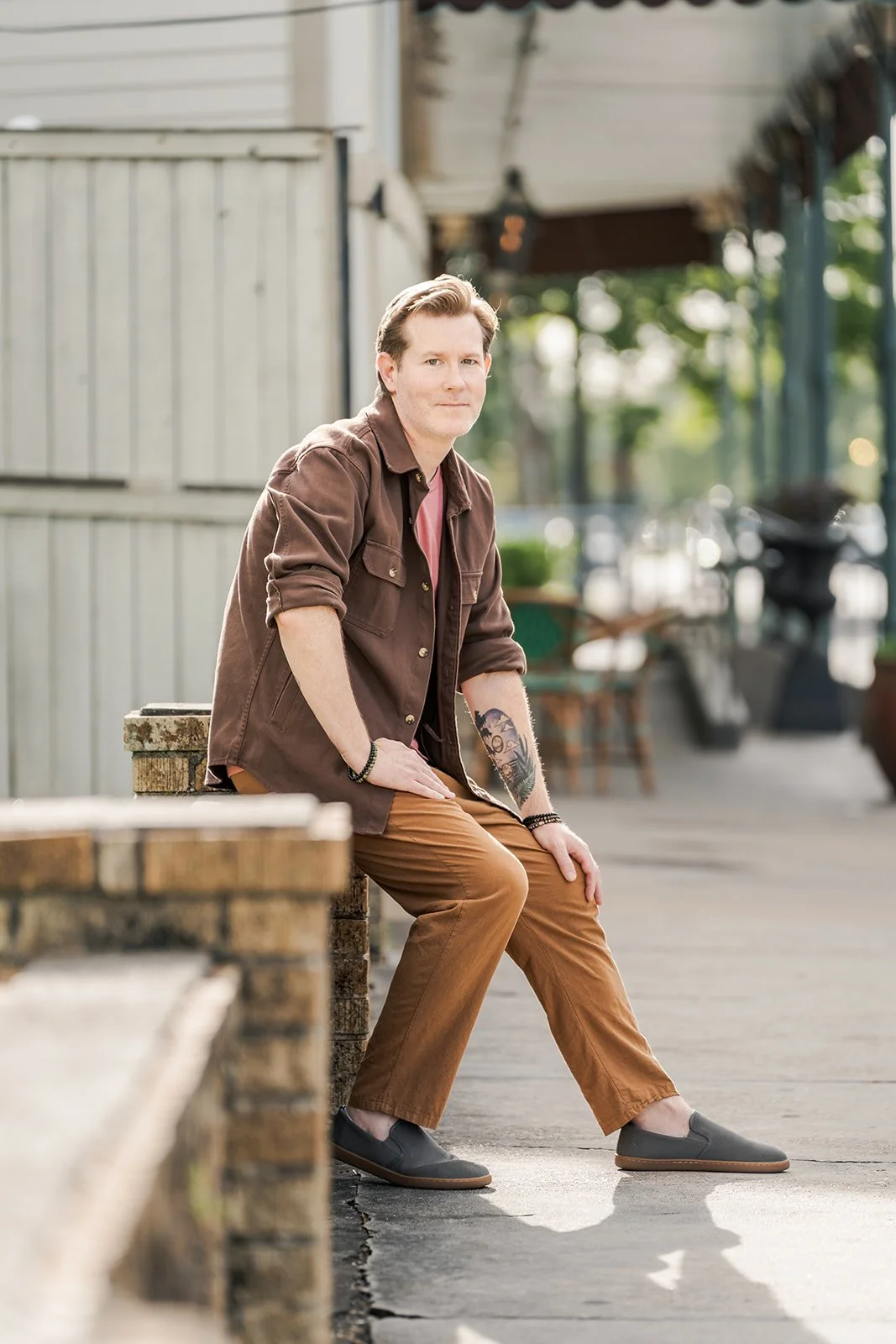 A young man with tattoos, wearing a brown jacket, pink shirt, and brown pants, sitting on a brick ledge outdoors in a park or town setting during daytime.