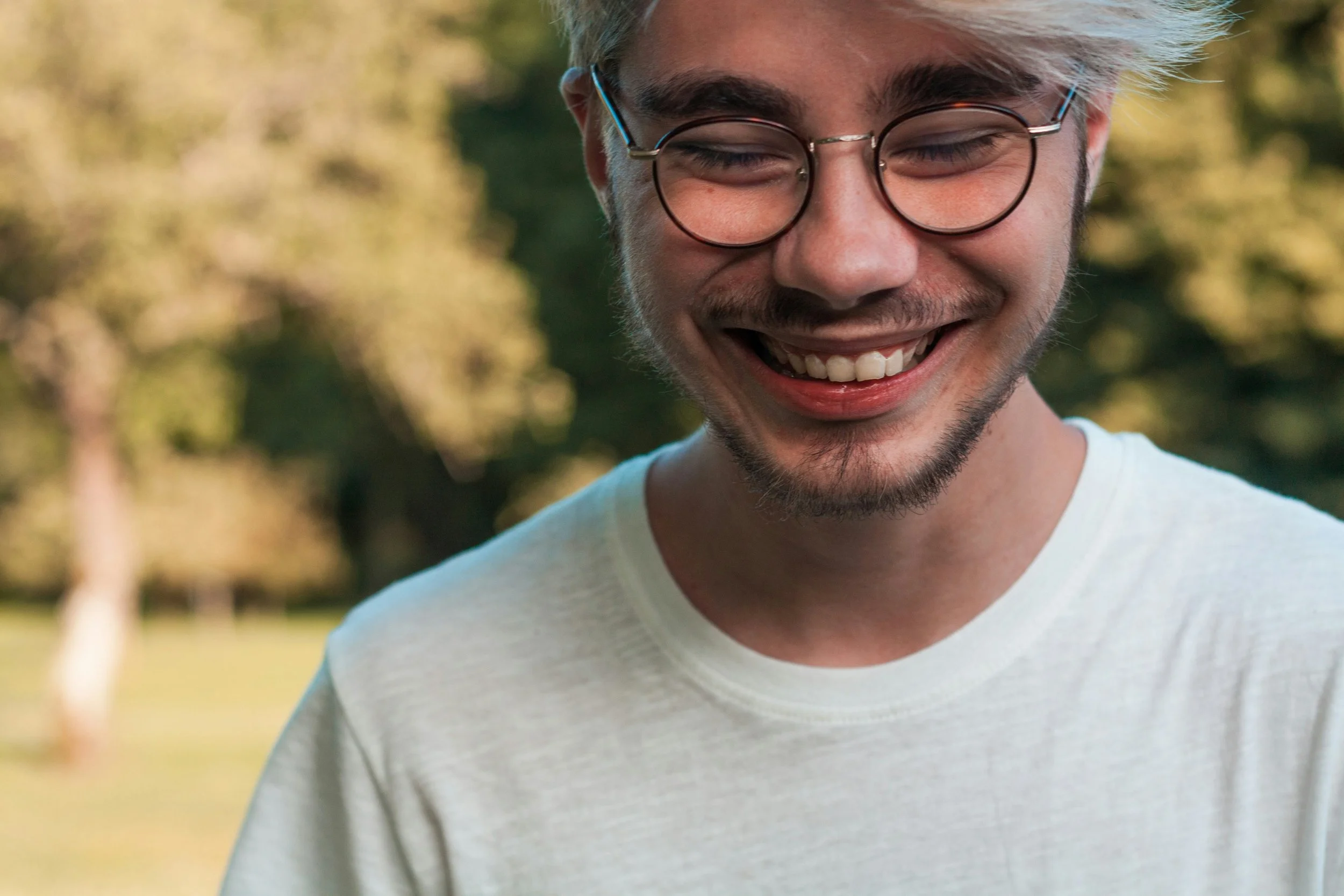 Close-up of a smiling young person with glasses and facial hair outdoors with blurred trees and grass in the background.