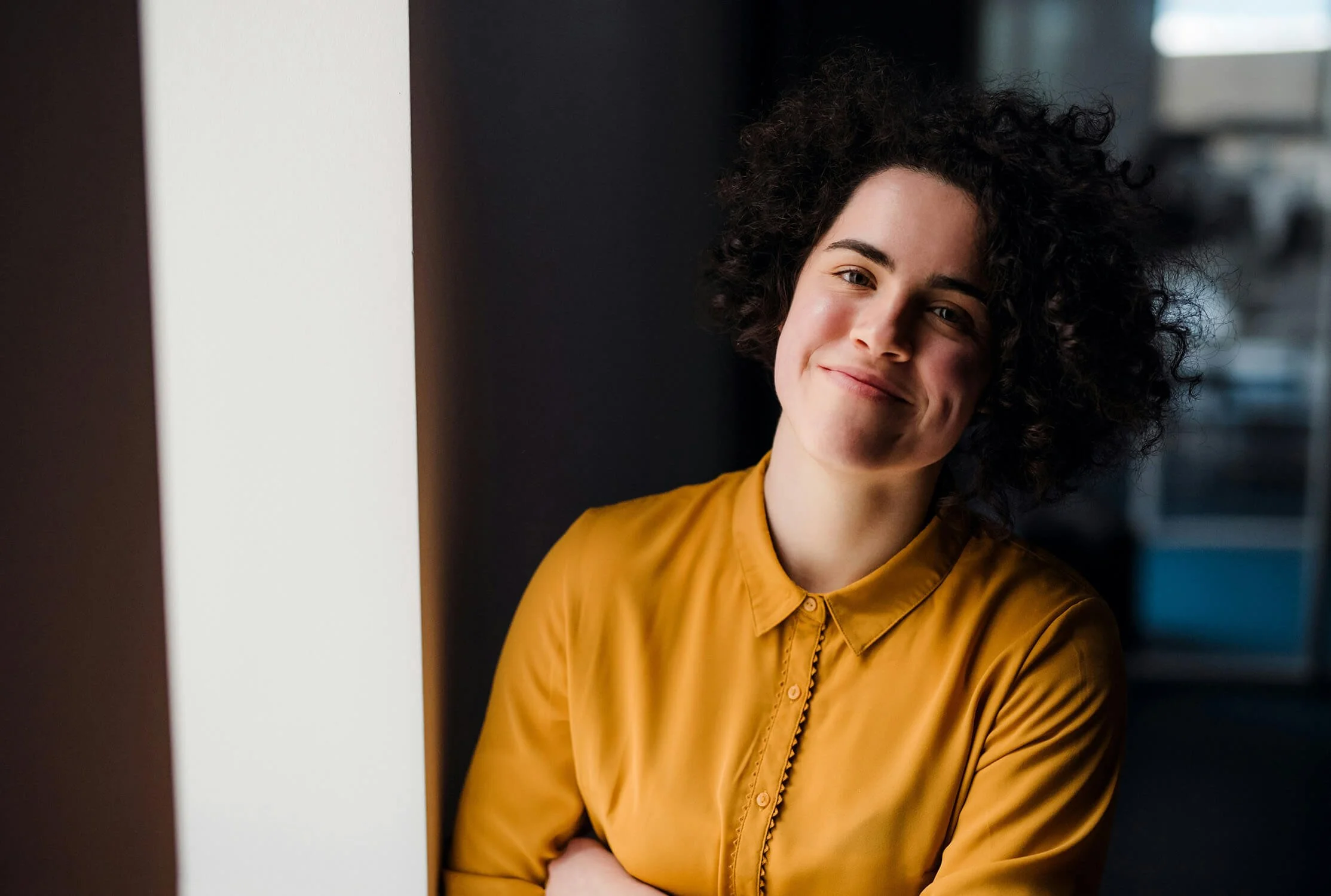 A woman with curly dark hair wearing a mustard yellow blouse, smiling and leaning against a white wall in an indoor setting.