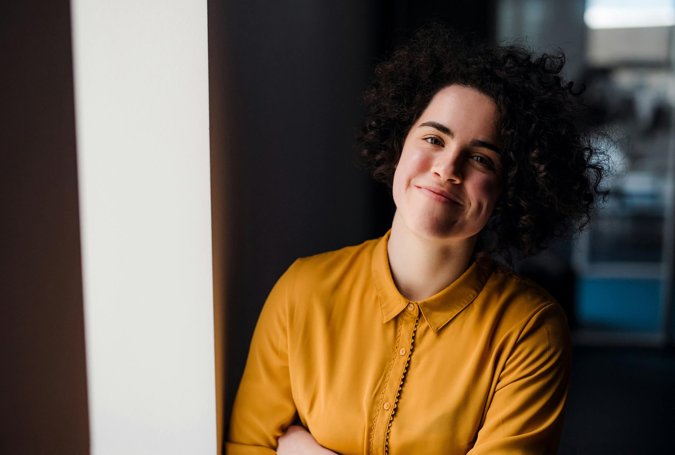 A woman with curly dark hair wearing a mustard yellow blouse, smiling and leaning against a white wall in an indoor setting.