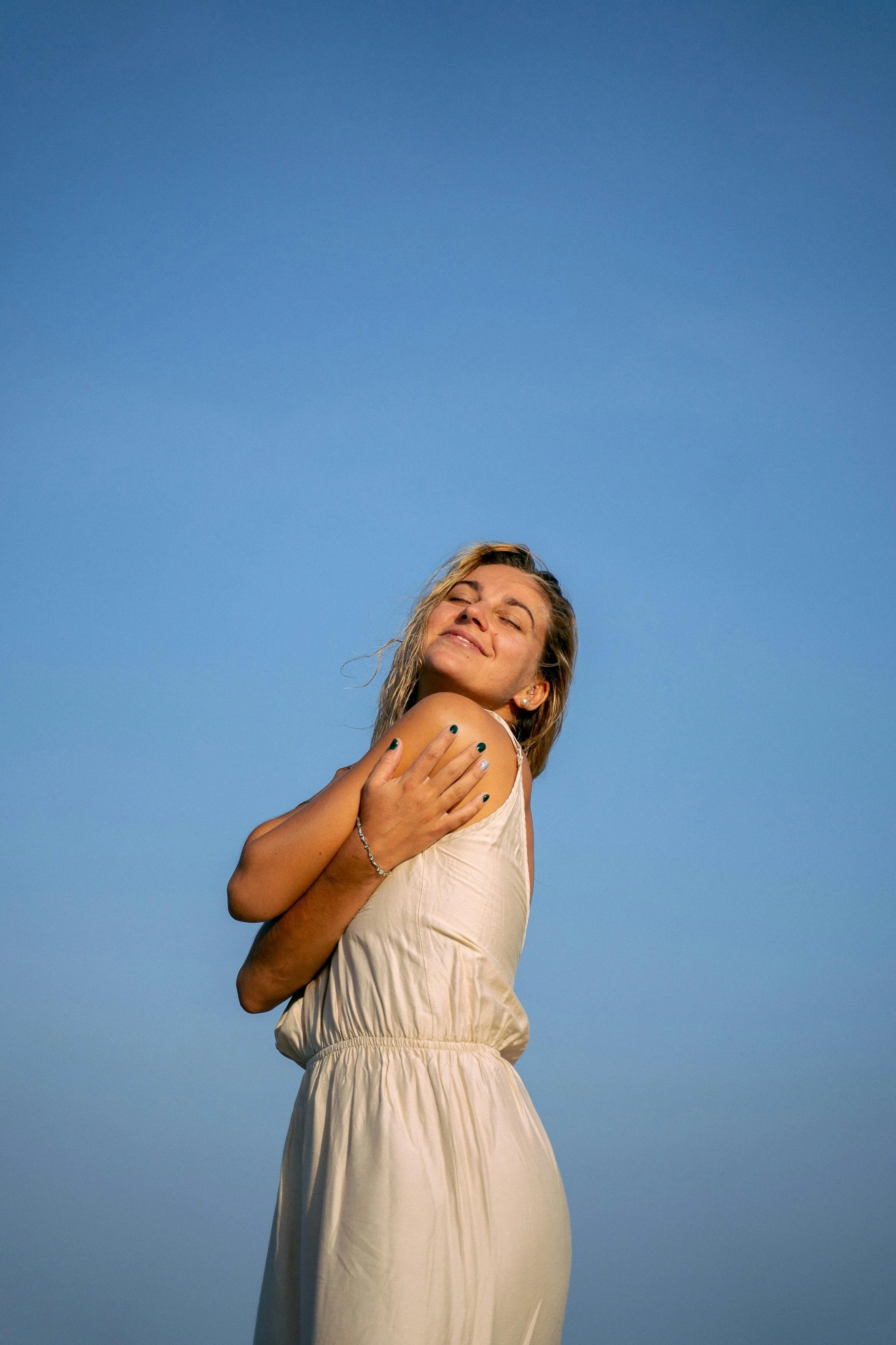 A woman with blonde hair smiling and hugging herself against a clear blue sky.