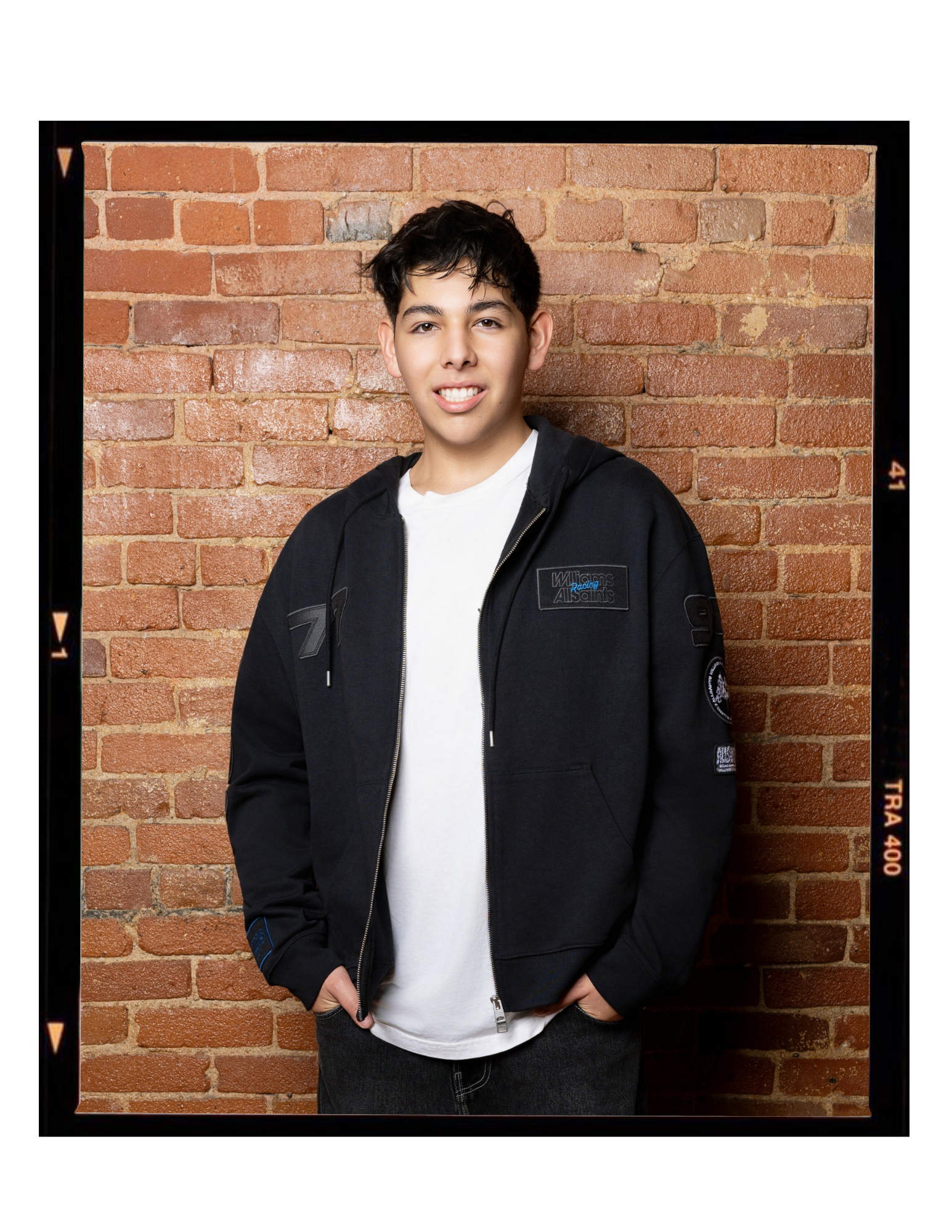 A young man with dark, curly hair standing in front of a red brick wall, wearing a black jacket with patches and a white t-shirt, smiling slightly with hands in pockets.