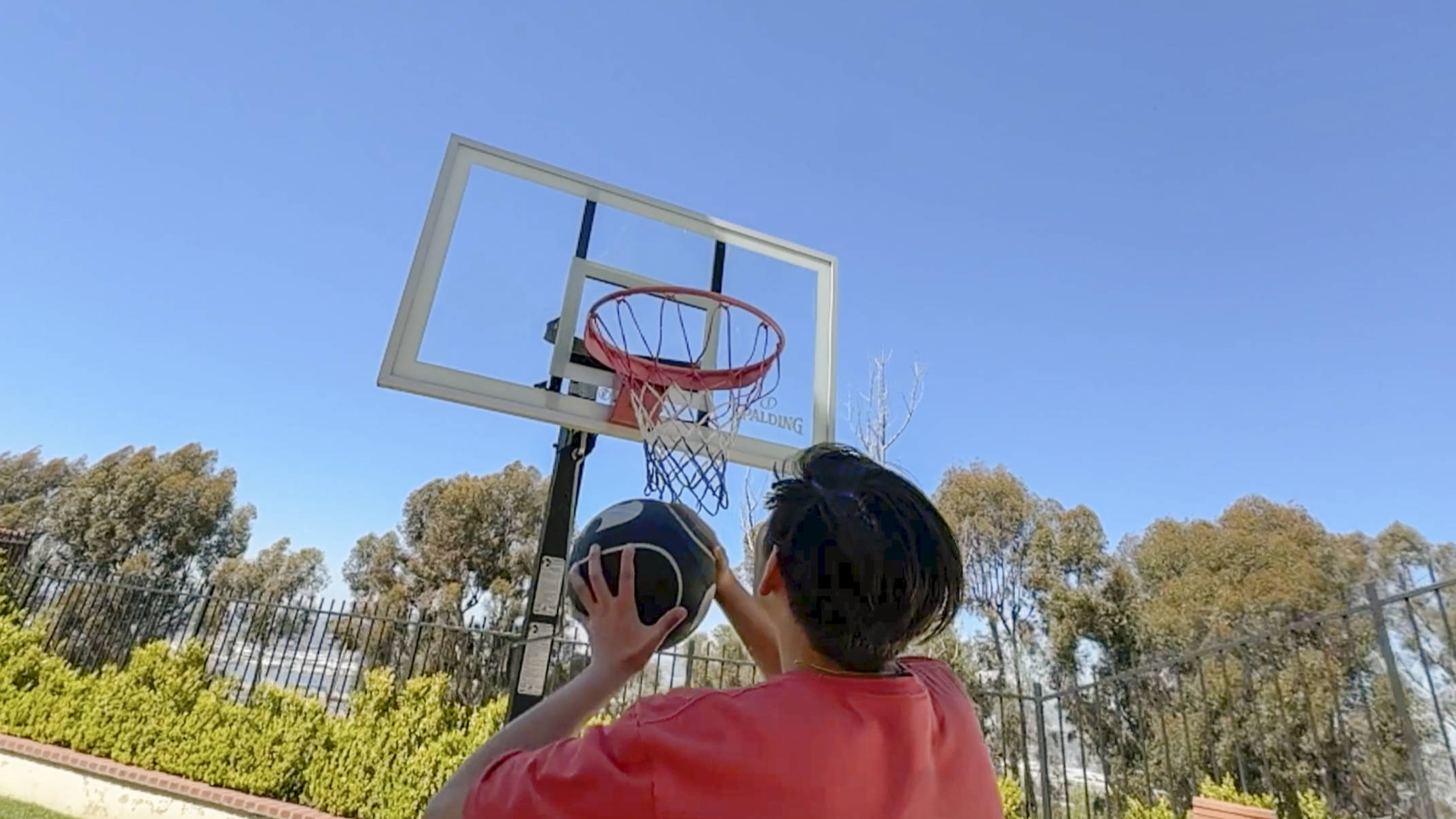 A young boy in a red shirt holding a basketball about to shoot at an outdoor basketball hoop under a clear blue sky.