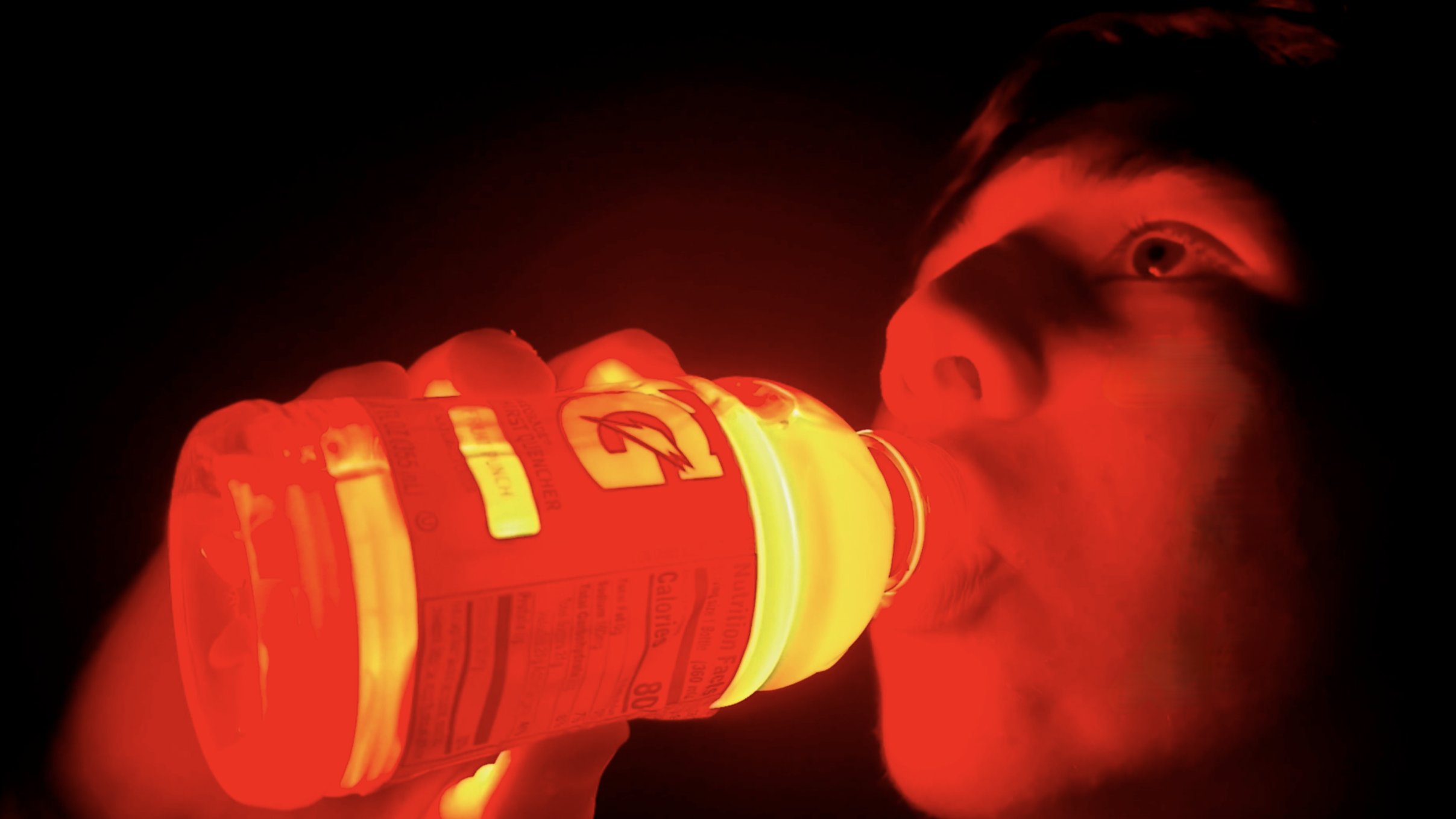 A person drinking from an illuminated bottle of an orange drink, with their face illuminated by the bottle's glow in a dark environment.