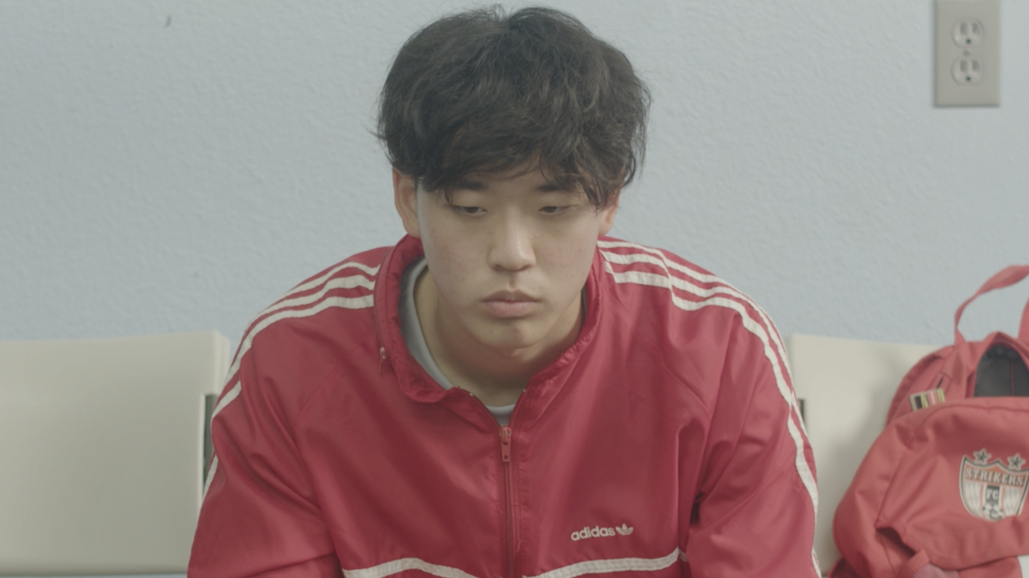 A young man with curly dark hair wearing a red Adidas jacket sitting in a room with a plain wall and electrical outlets visible behind him.