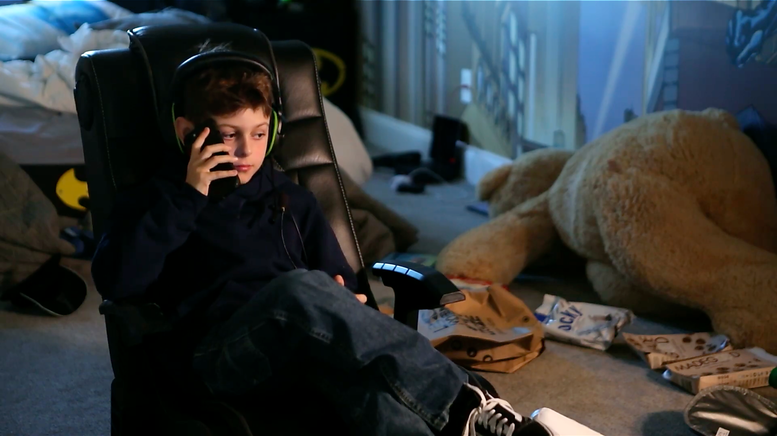 A young boy sitting in a black gaming chair, wearing headphones, talking on a phone, with snacks and packages in front of him in a cluttered room.