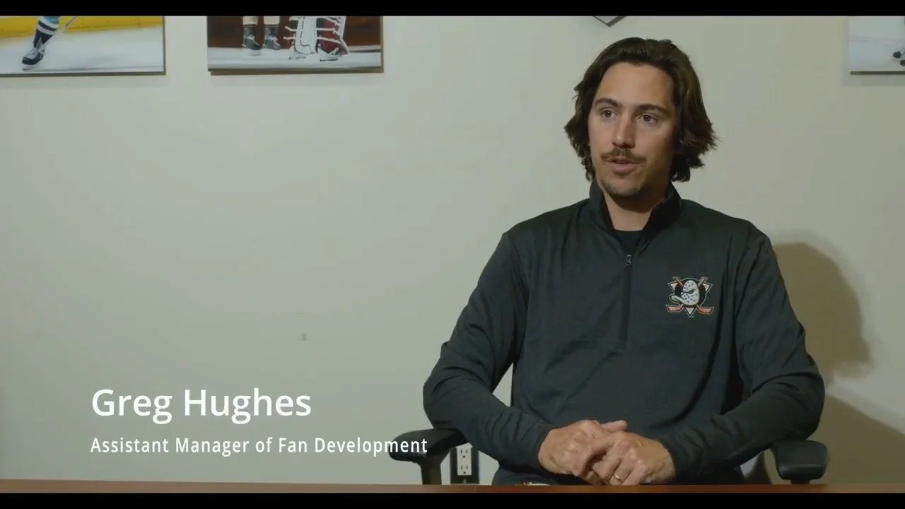A man with shoulder-length brown hair and a mustache sitting at a desk, wearing a black jacket with a hockey team logo. Behind him is a wall with hockey-themed photos. The text on the image reads "Greg Hughes, Assistant Manager of Fan Development."