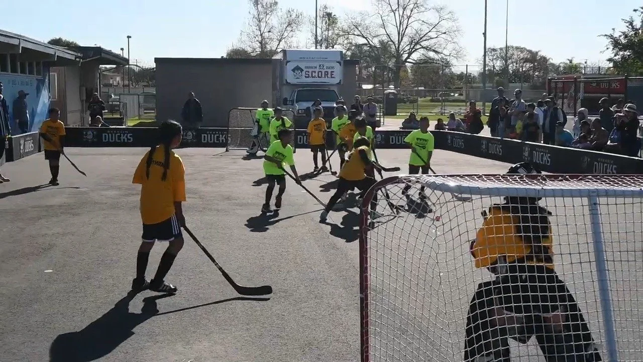 Hockey game with young players on outdoor rink, goalie in net, spectators watching, sunny day.