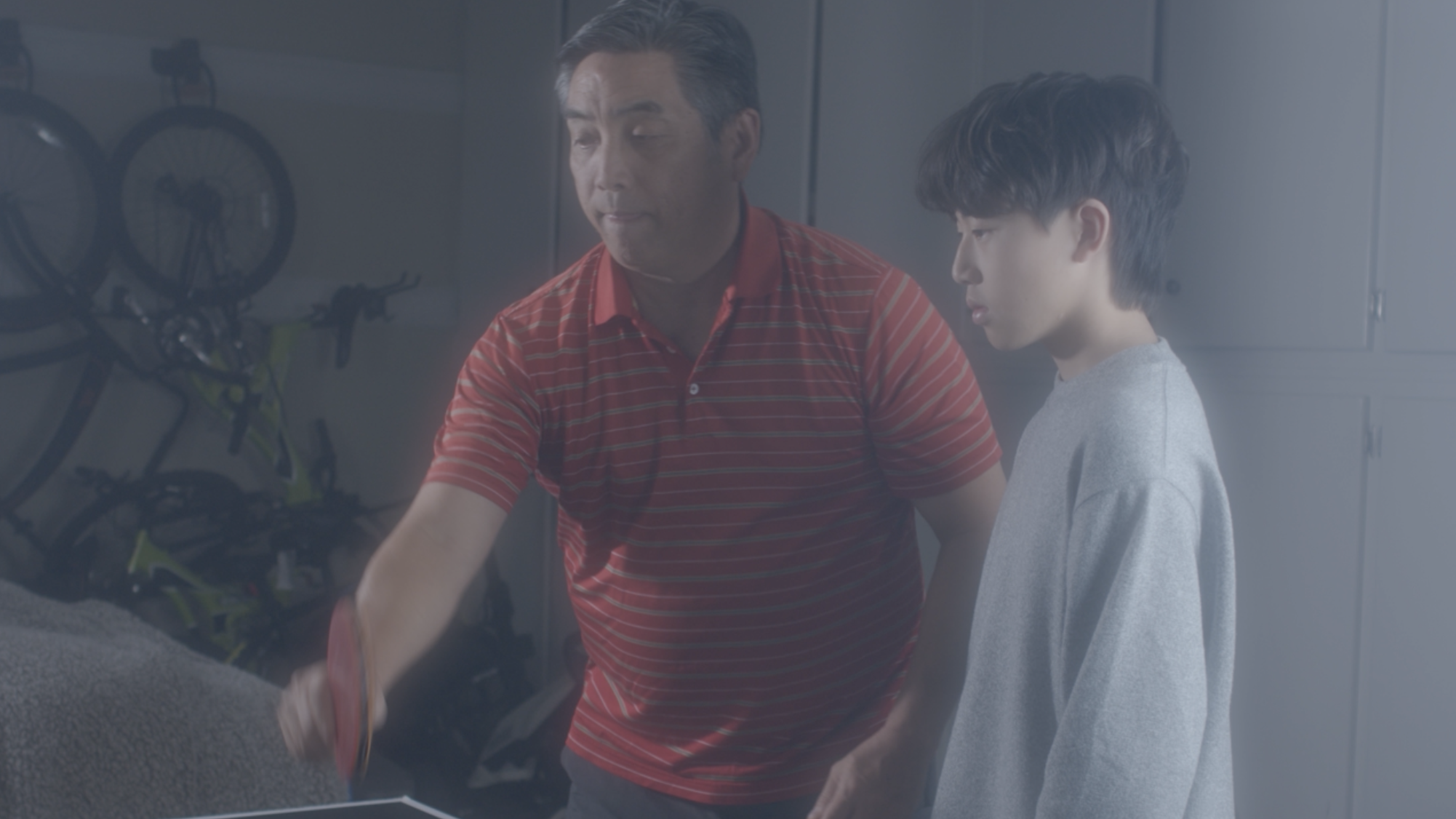 An adult man in a red polo shirt with a younger man with dark hair holding a table tennis paddle, in a garage.