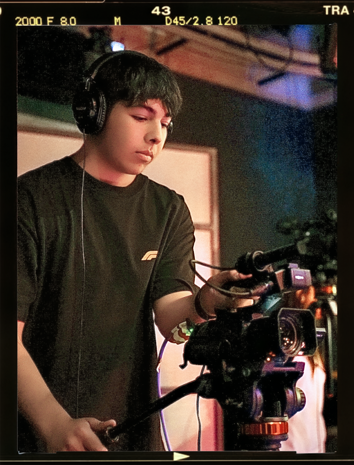 A young man with dark hair wearing headphones operating a professional video camera on a film set.