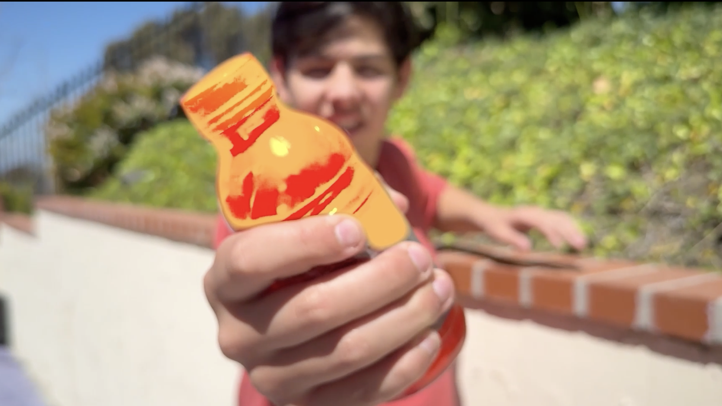 A young person with dark hair smiling and holding a bright orange and red bottle close to the camera outdoors.