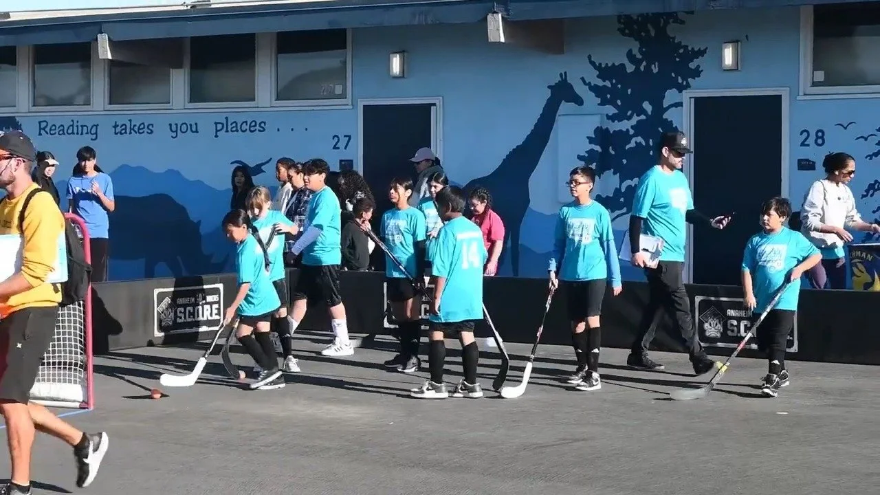 Young hockey players in blue jerseys and black shorts practicing on a parking lot with a goal, outside a building with a mural of a giraffe, trees, and mountains, with a quote reading "Reading takes you places..."
