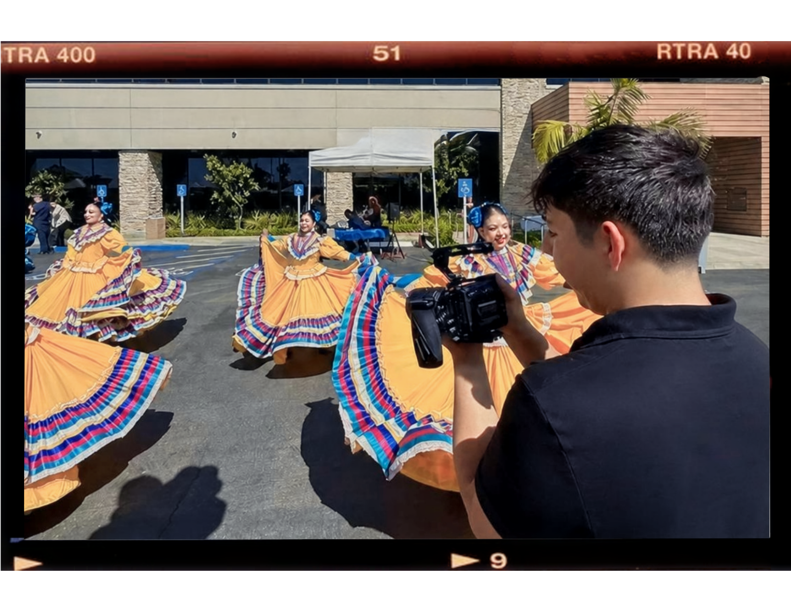 A man filming a dance performance of women in bright yellow mexican ballet folklorico traditional dresses dancing outdoors on a sunny day.