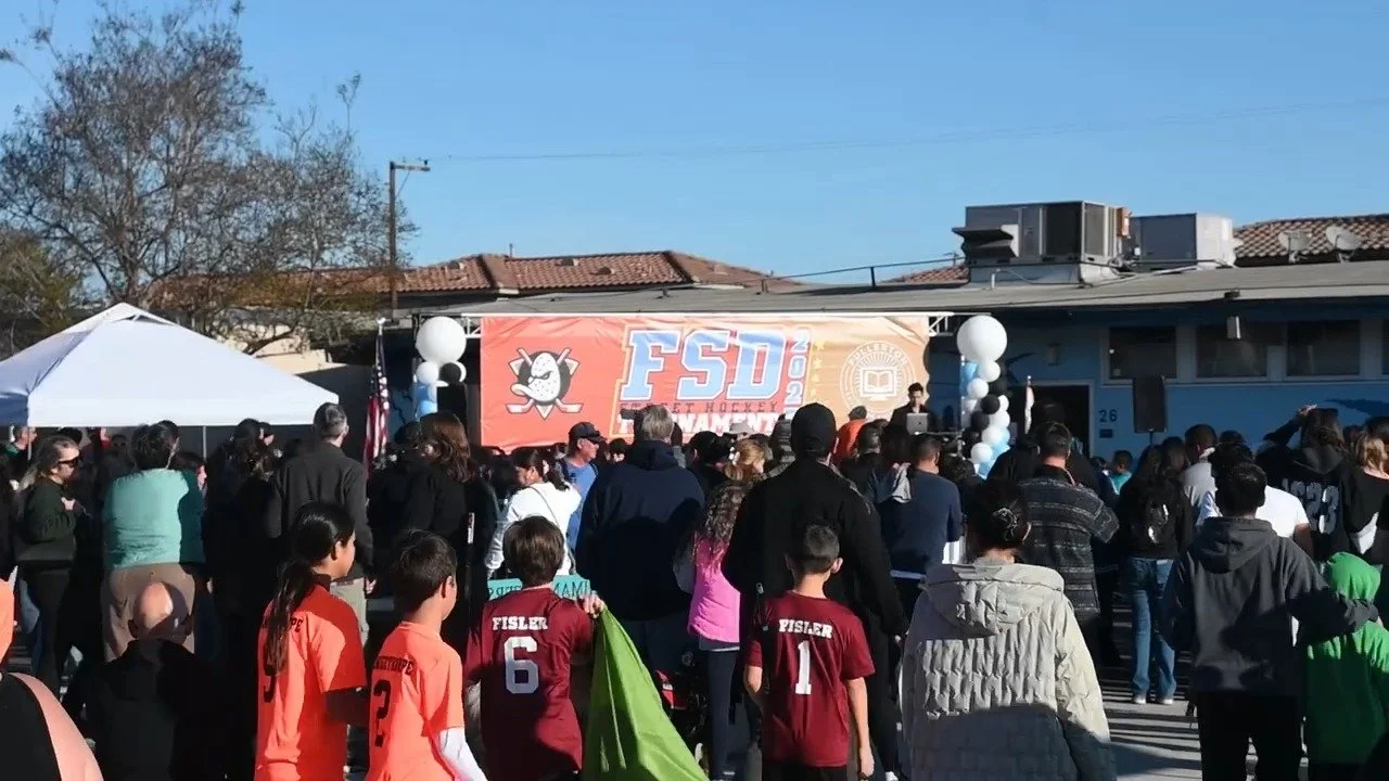 Crowd of people gathered outside for an event with a large banner reading 'FSD' and hockey-themed graphics, on a sunny day.