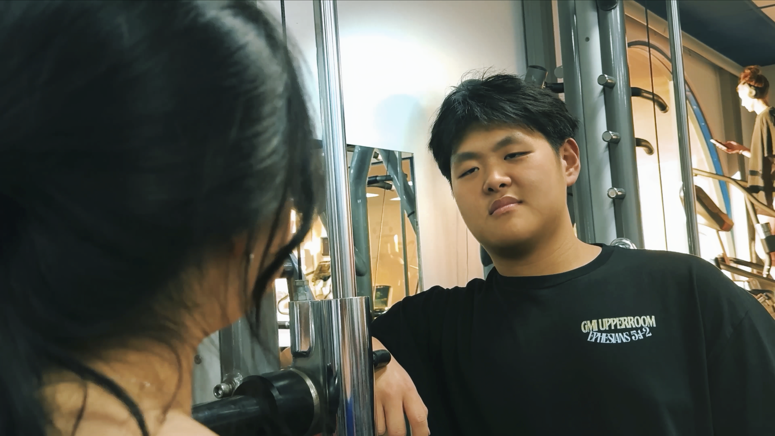 A young man with short black hair and a black T-shirt with white text is inside a gym, looking at a woman with dark hair, who is partially visible in the foreground, also in the gym. 