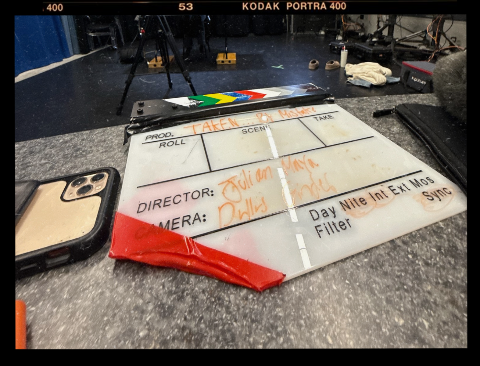 A film slate with handwritten notes and commands, situated on a gray speckled surface. The background shows various behind-the-scenes film equipment.