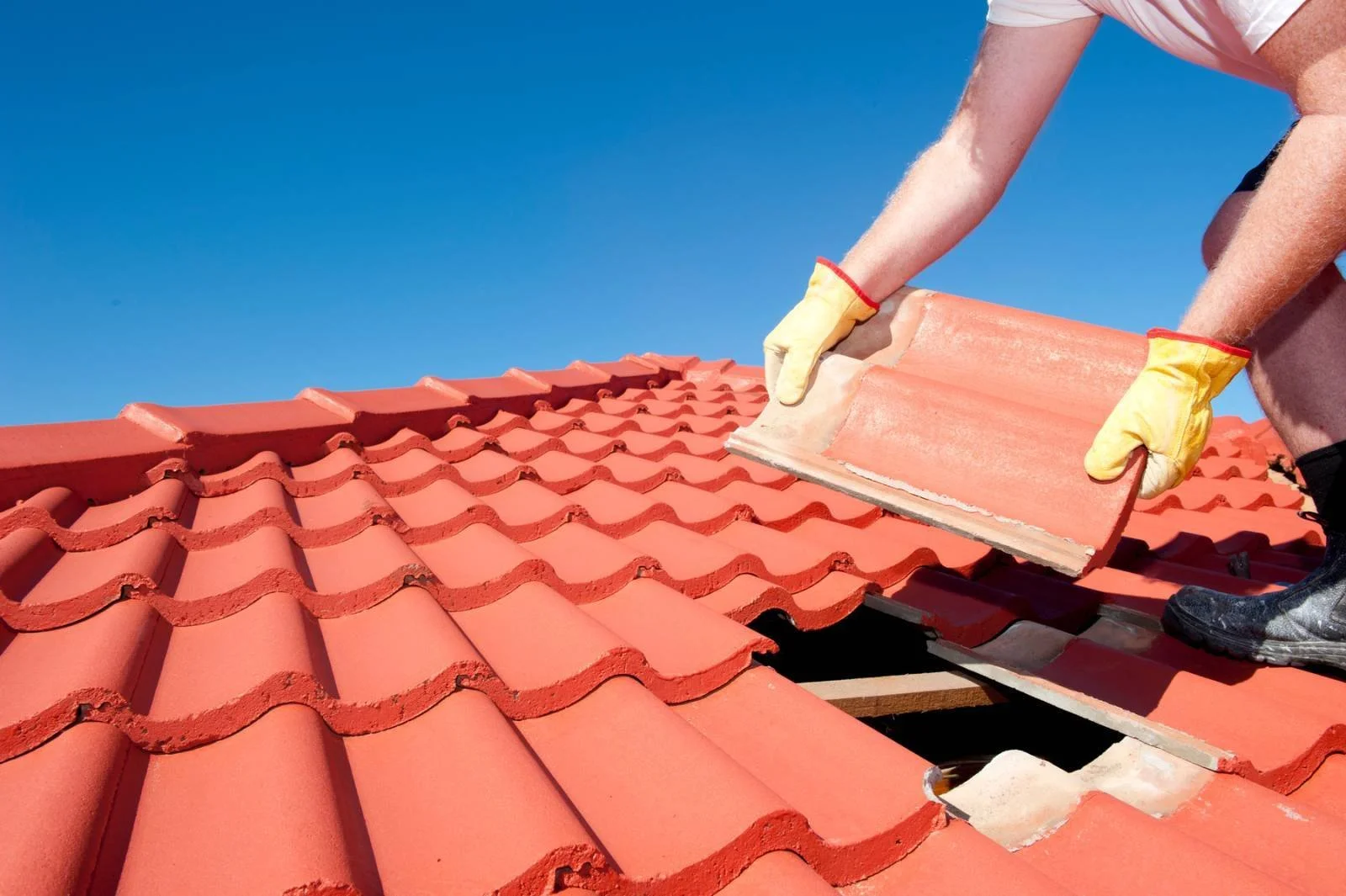 Travaux de pose de tuiles rouges sur un toit, personne en train de placer une tuile, ciel bleu en arrière-plan.