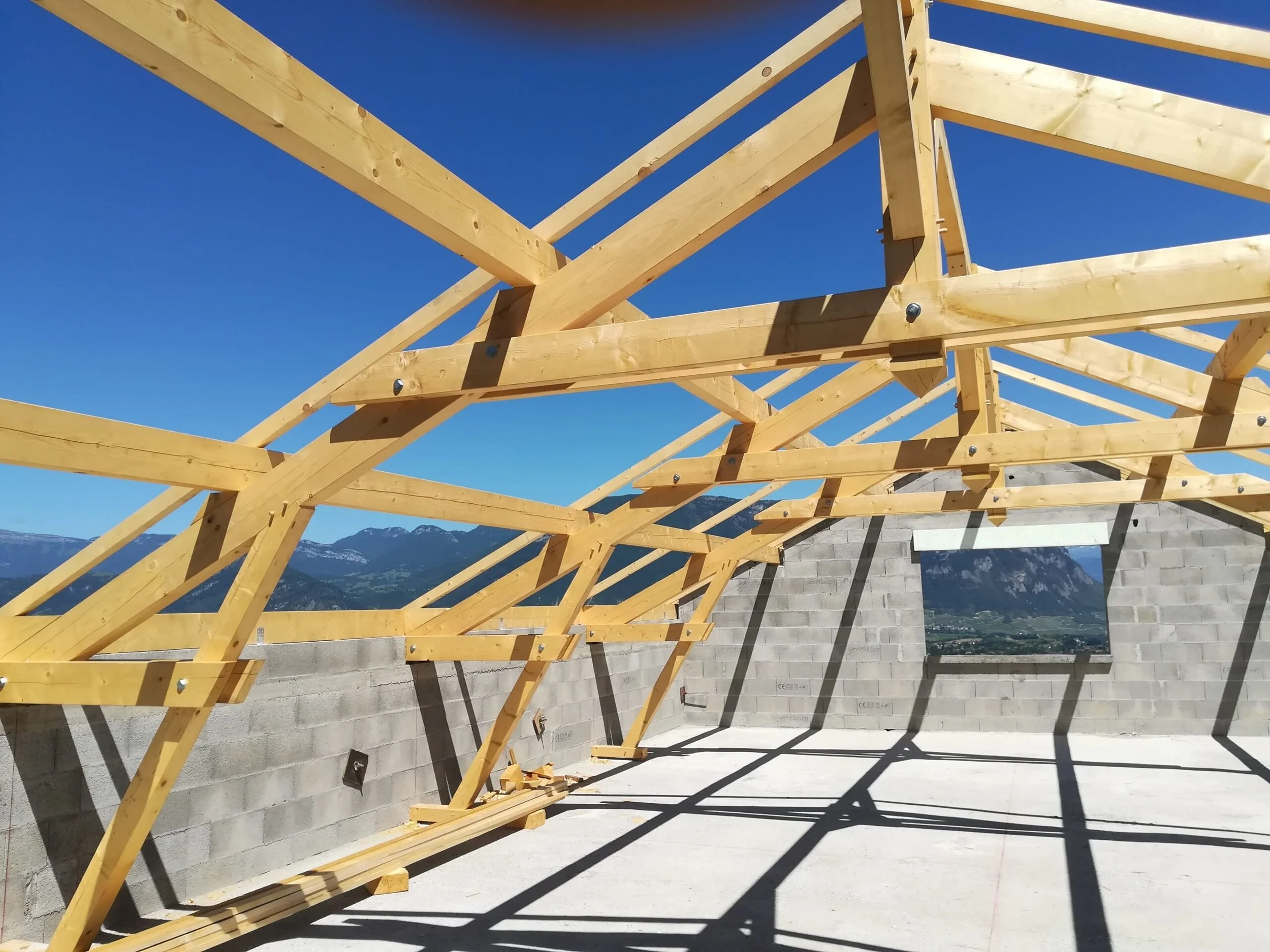 Structure en bois en construction avec des poutres et des supports en bois contre un ciel bleu et des montagnes en arrière-plan.