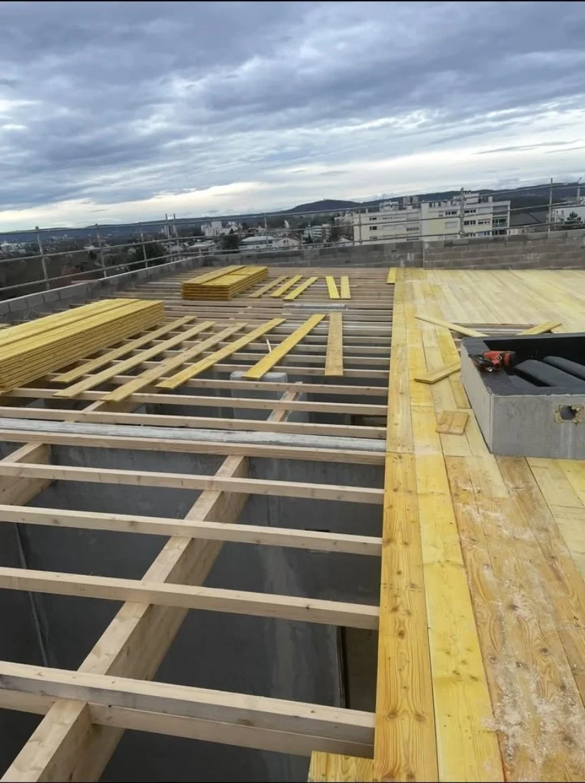 Travaux de construction d'une toiture avec bois, planches en cours de pose, nuages dans le ciel, bâtiments et horizon en arrière-plan.
