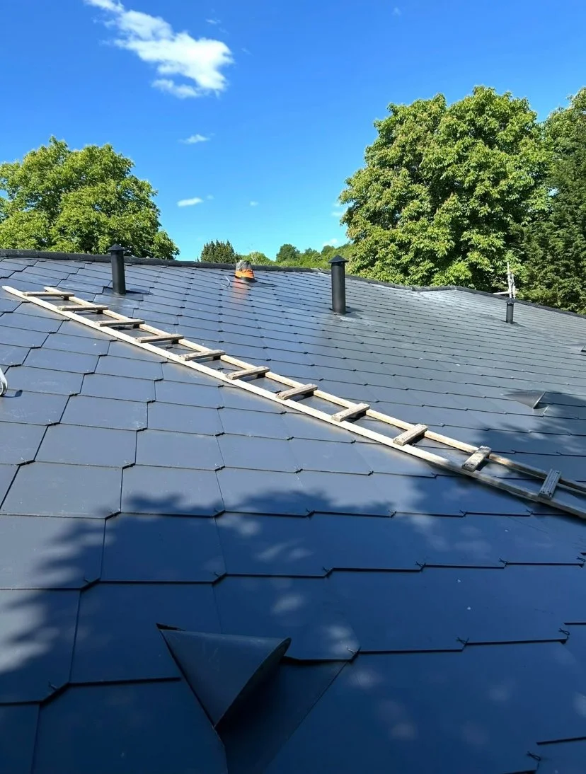 Toit en tuiles noires avec une échelle en bois et plusieurs conduits de cheminée, sous un ciel bleu avec quelques nuages, entouré d'arbres verts.