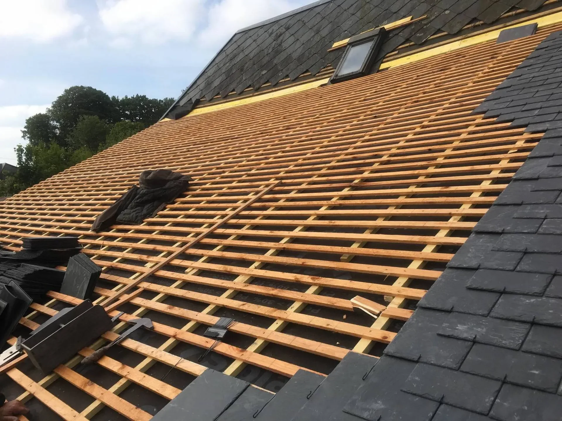 Toit en cours de rénovation avec structure en bois, quelques tuiles noires posées, une fenêtre de toit et des outils sur le toit.