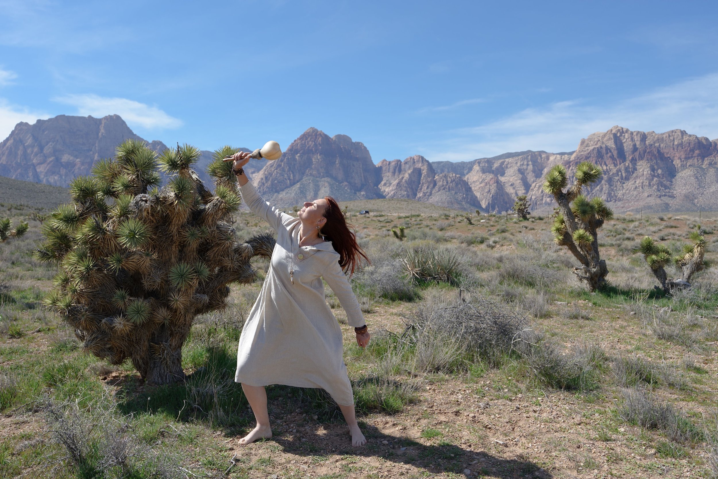 A woman dressed in a white coat standing barefoot in a desert landscape with Joshua trees, holding a large bone with a ball on top, with mountains in the background and a clear blue sky.