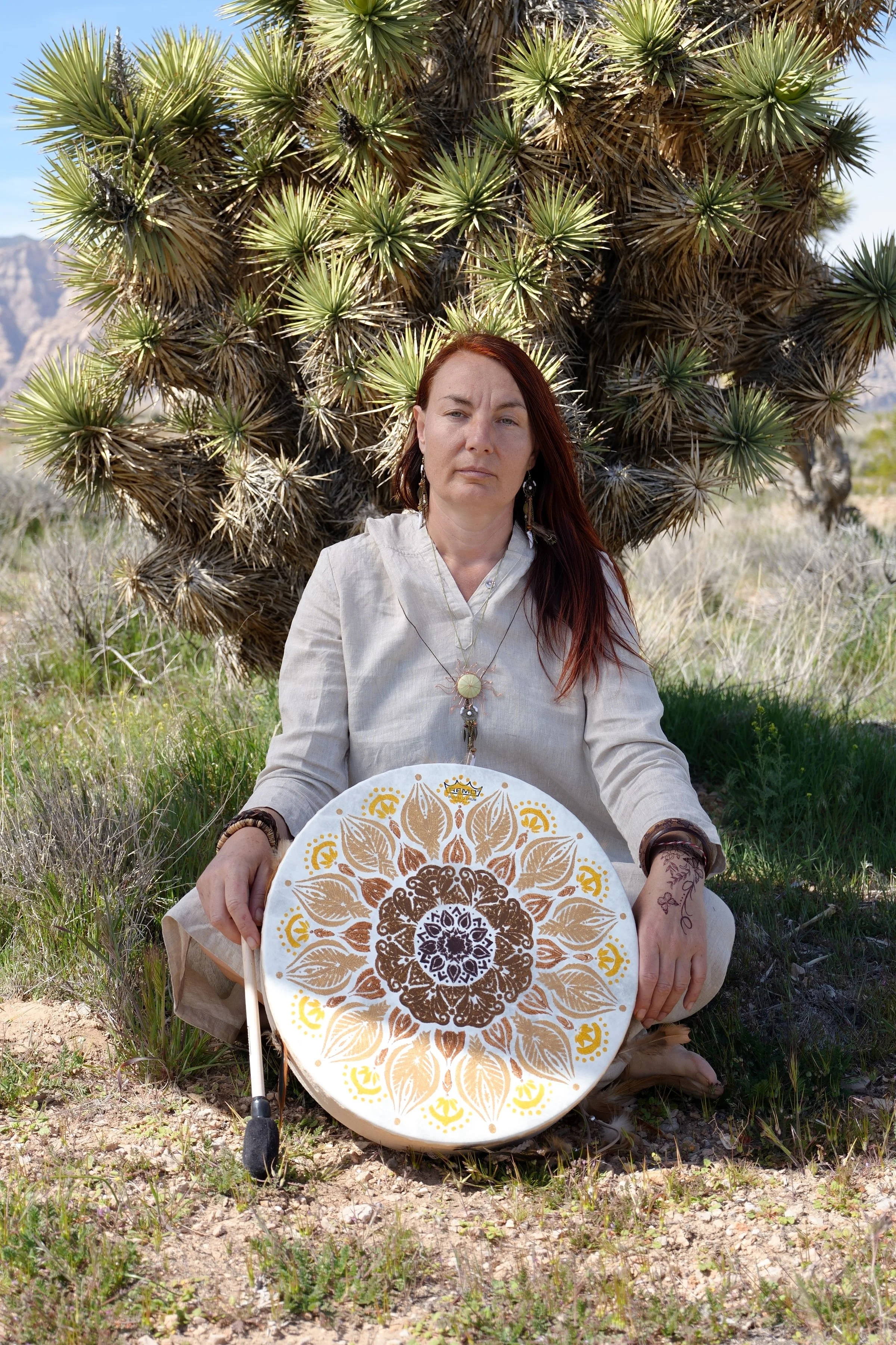 A woman with long red hair and tattoos on her hand, sitting on the ground in front of a large desert cactus, holding a round decorative object with an intricate mandala pattern in brown, yellow, and white.