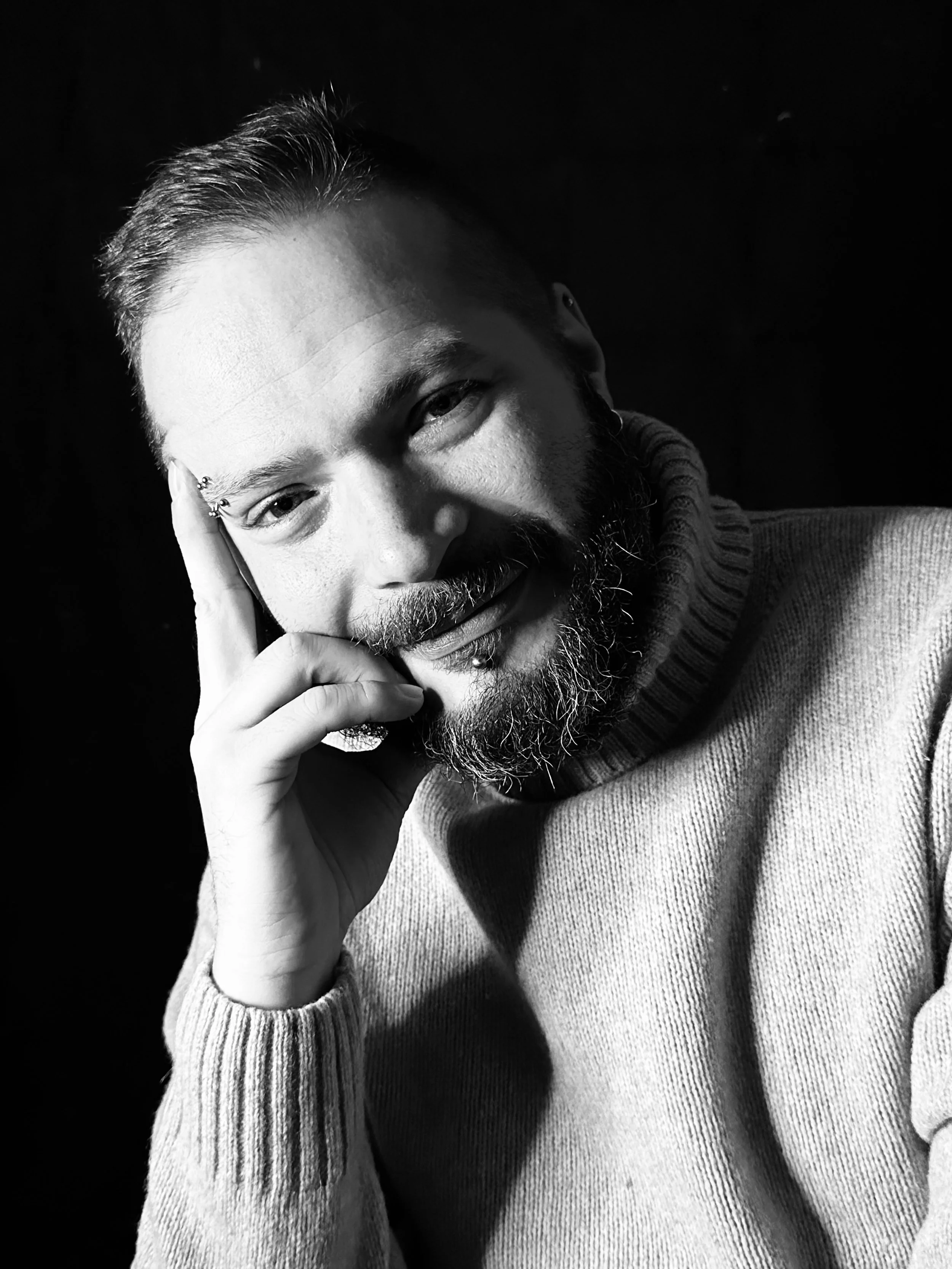 Black and white portrait of a man smiling, with his hand resting on his face, wearing a turtleneck sweater.