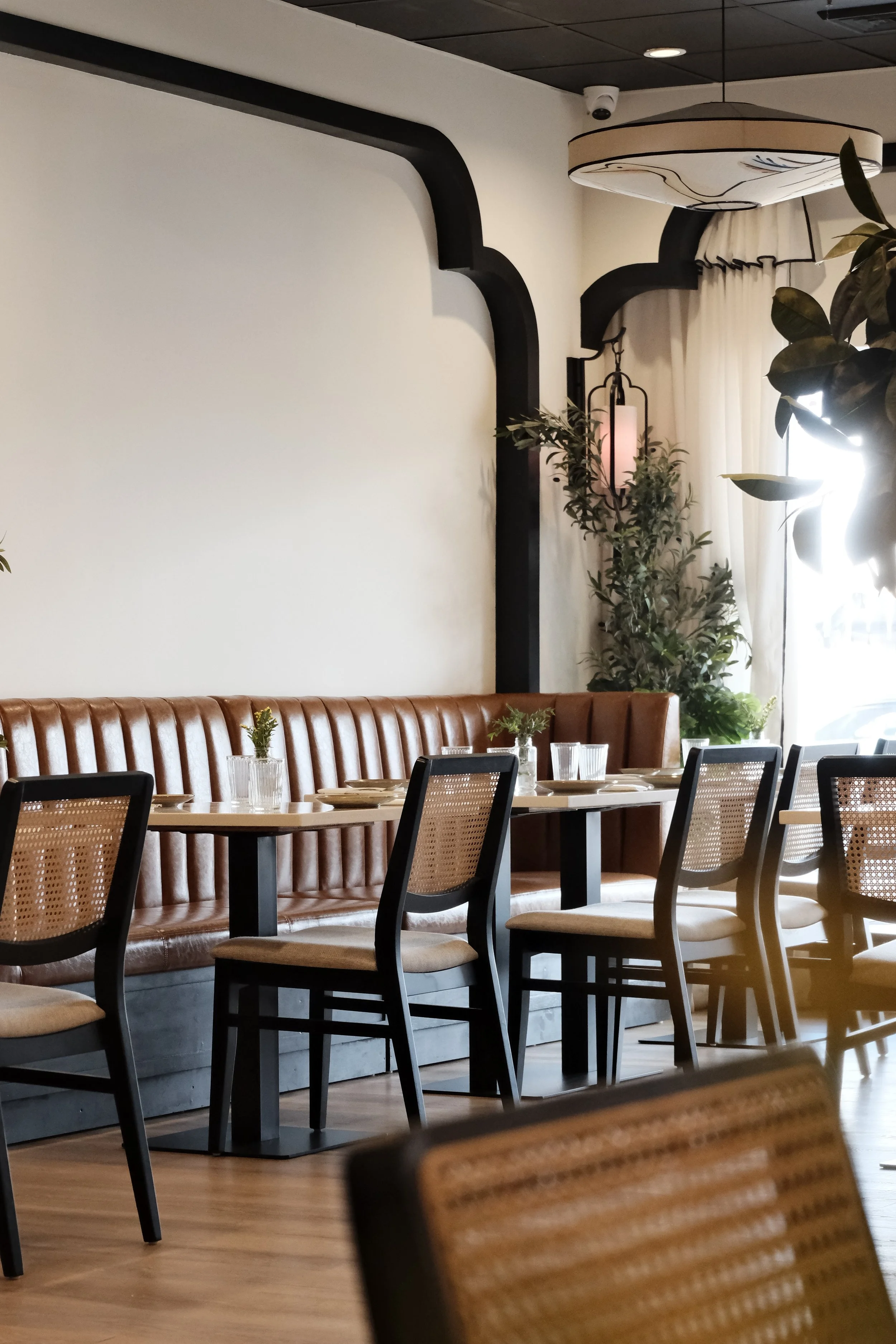 Interior of a restaurant with wooden tables, cushioned chairs, a leather booth, large plants, hanging lanterns, white curtains, and a white wall with decorative black accents.