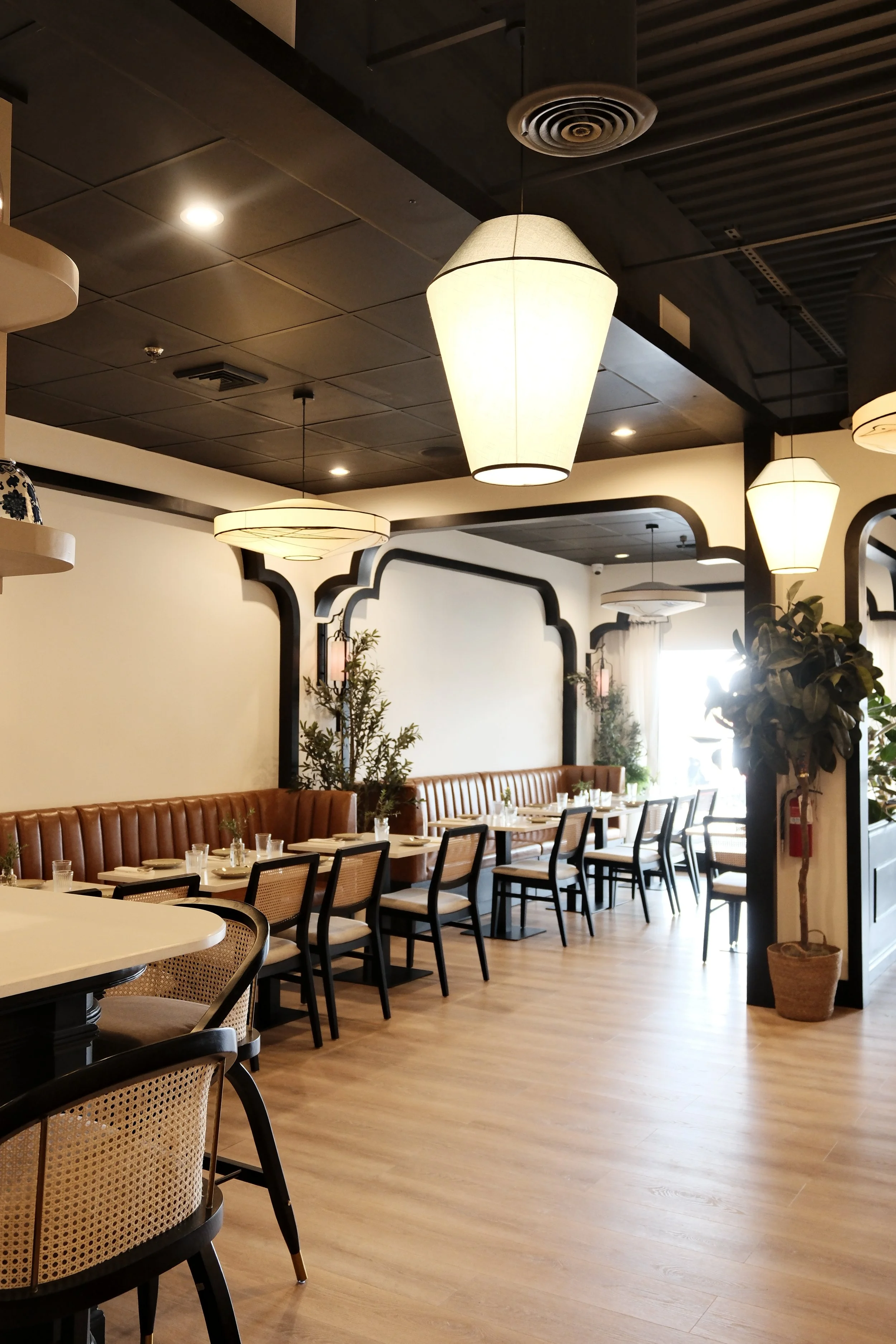 Upscale restaurant interior with hanging lamps, tables set with white tableware, chairs, and potted plants, featuring wooden floors and decorative wall accents.