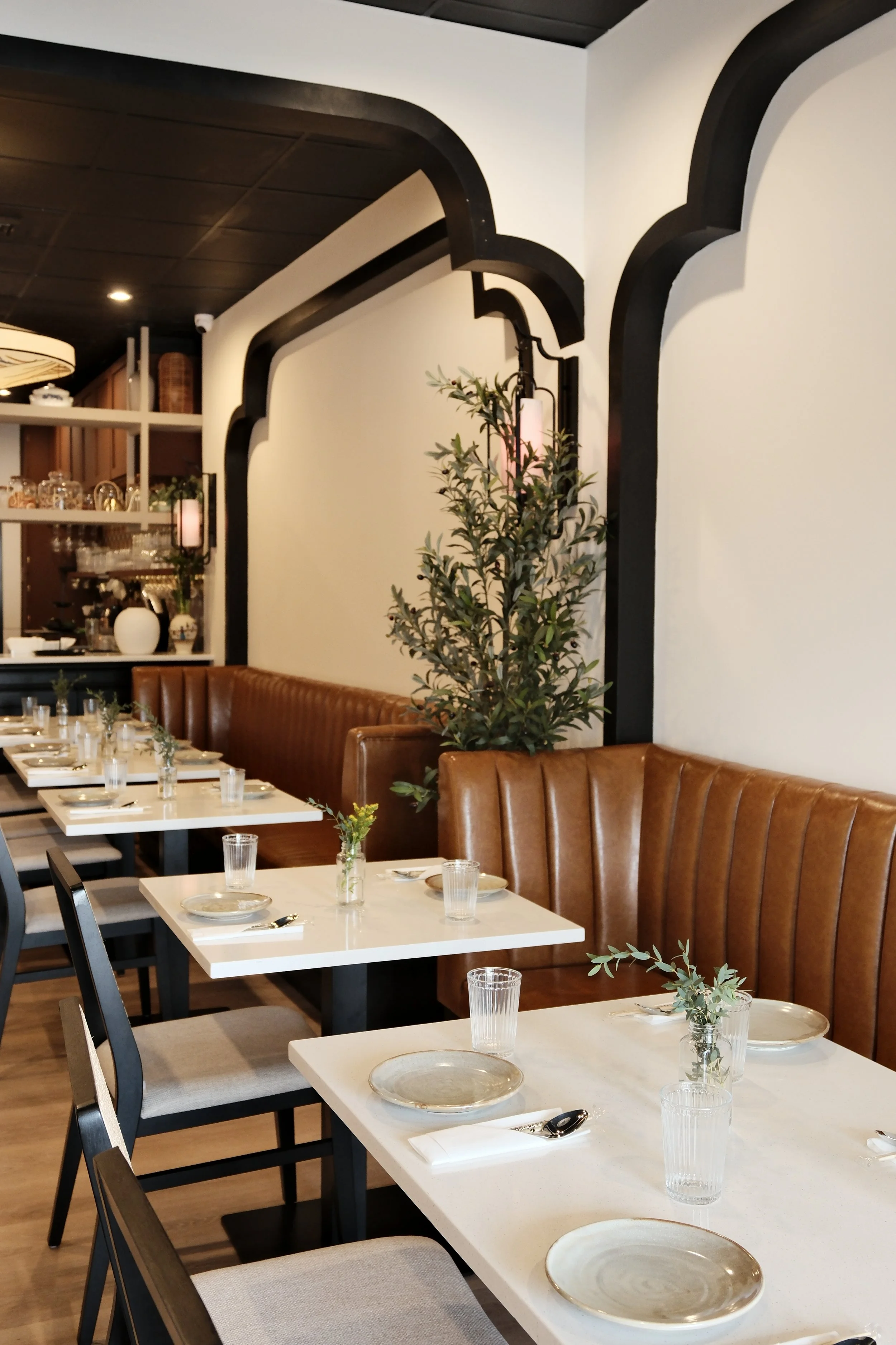 A row of white tables set with plates, glasses, and silverware in a restaurant. Brown leather chairs line the tables, and a large green potted plant is positioned beside a decorative wall with black trim and white walls. In the background, shelves hold glassware and decorative items.