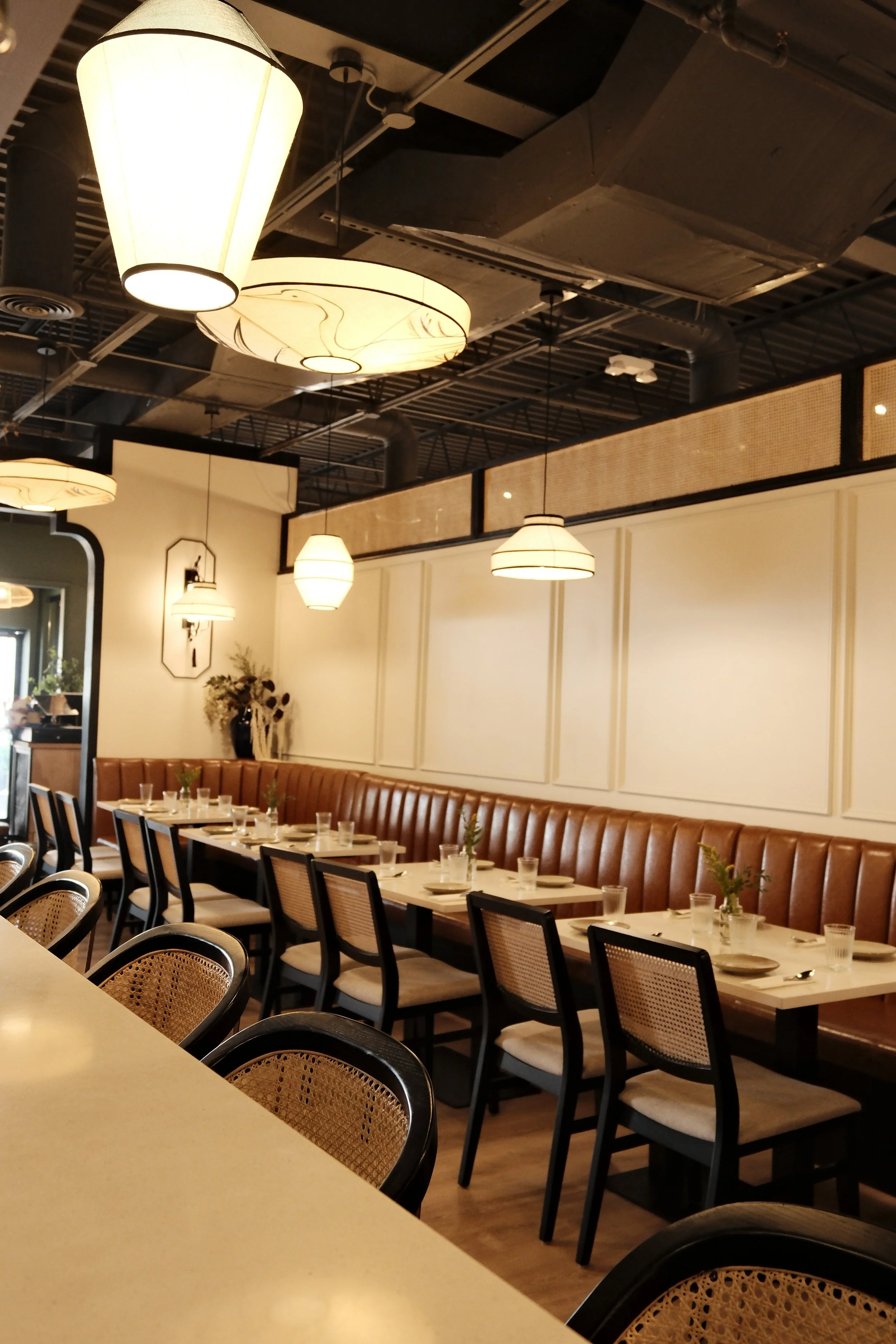 Interior of a restaurant with a long row of tables set with glasses, plates, and utensils, and a brown leather bench along the wall. Decorative lighting fixtures hang from the ceiling, emitting warm light. The wall has beige paneling, and there are plants and wall art visible.