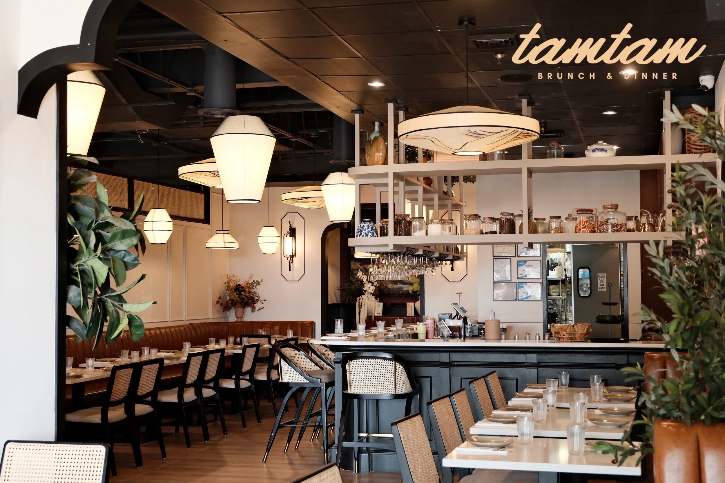 Interior of a restaurant named Tamaram with wooden ceiling, hanging lamps, tables set with glasses and plates, a shelf with jars, and greenery.