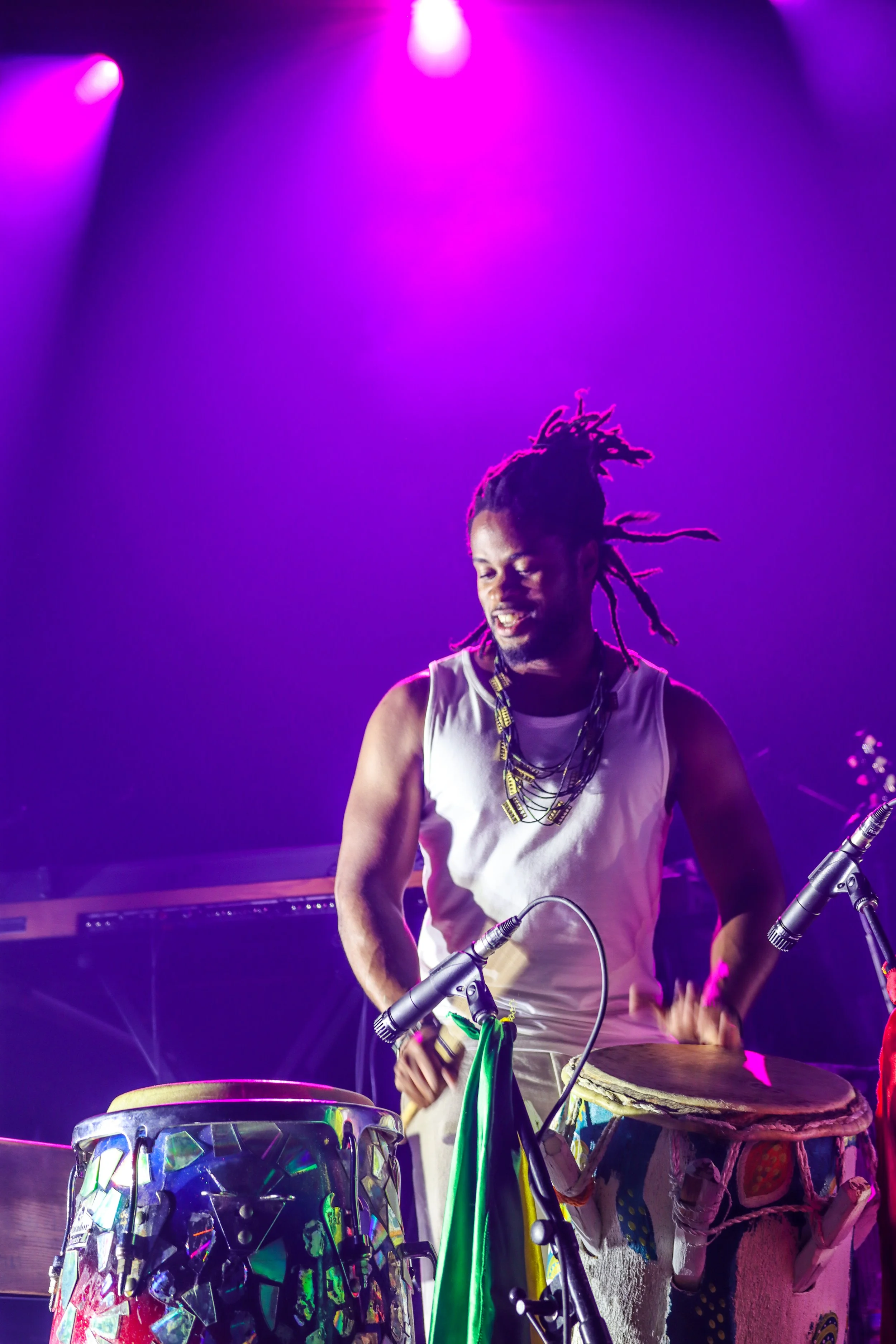 A Haitian musician with dreadlocks playing drums and percussion instruments on stage with arcade fire and boukman eksperyans, illuminated by purple and pink stage lights.