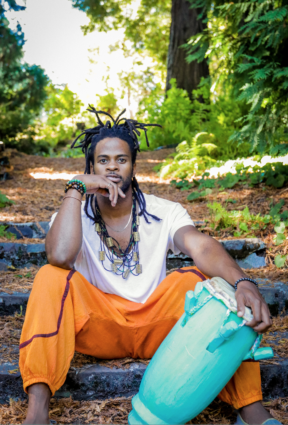 A man with dreadlocks sitting outdoors on a stone step, holding a large drum, surrounded by green foliage and trees.