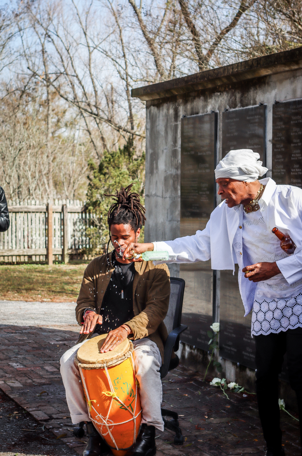 A man with dreadlocks playing a drum is being sprayed with a liquid by an older woman dressed in white, standing outdoors near a memorial wall.