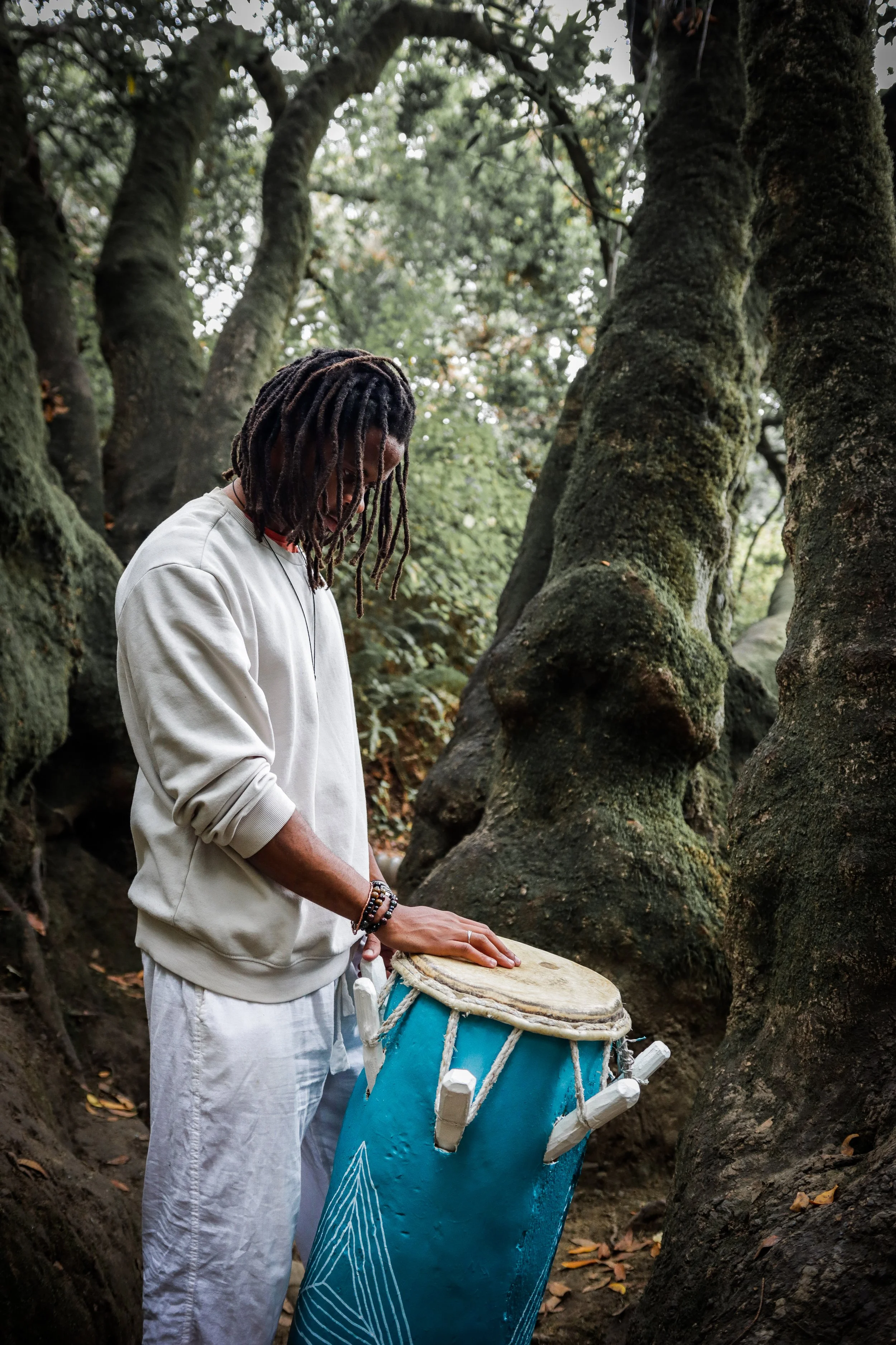 A person with dreadlocks wearing a white sweatshirt and white pants, standing in a forest, playing a drum with a blue body and white top.