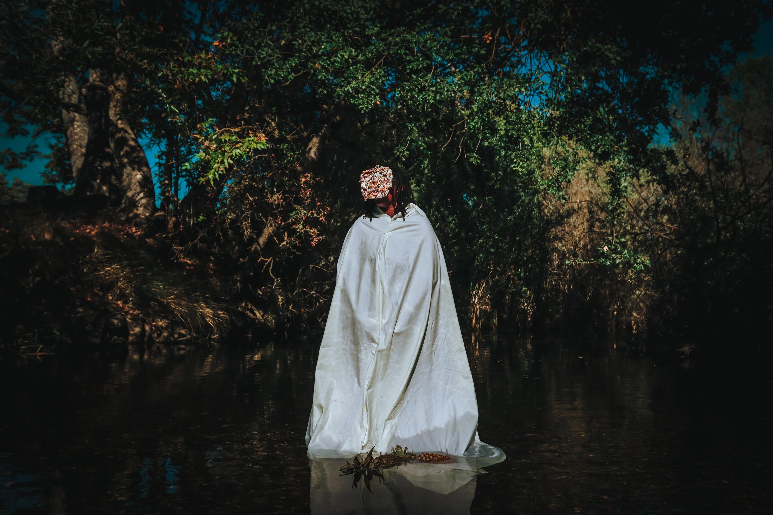 A person standing in a body of water at night, wearing a white cloak, with a large bouquet of flowers covering their face, surrounded by dark trees and a clear blue sky reflected in the water.