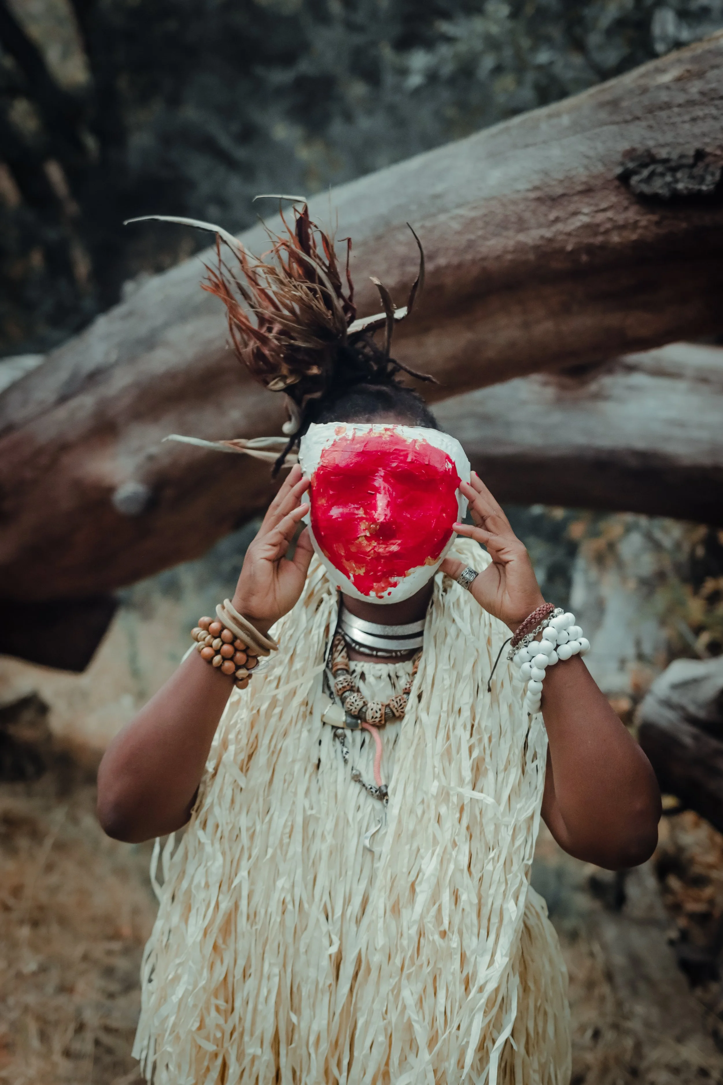 Person wearing traditional attire and a mask, holding a red and white painted object, outdoors with a large fallen tree in the background.