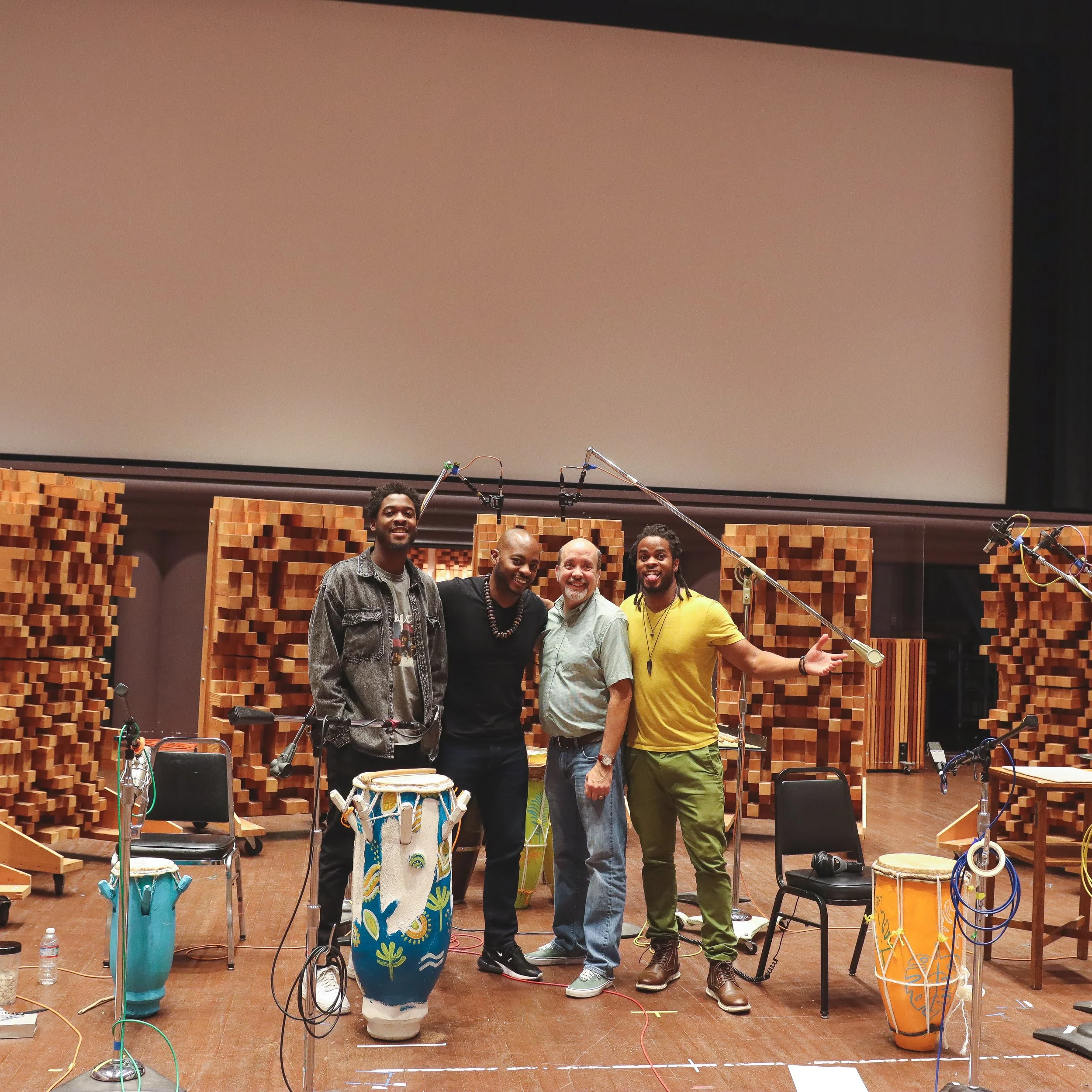 Group of five men standing together in a music recording studio, surrounded by sound-absorbing wooden panels, microphones, and musical equipment, smiling and posing for the photo.